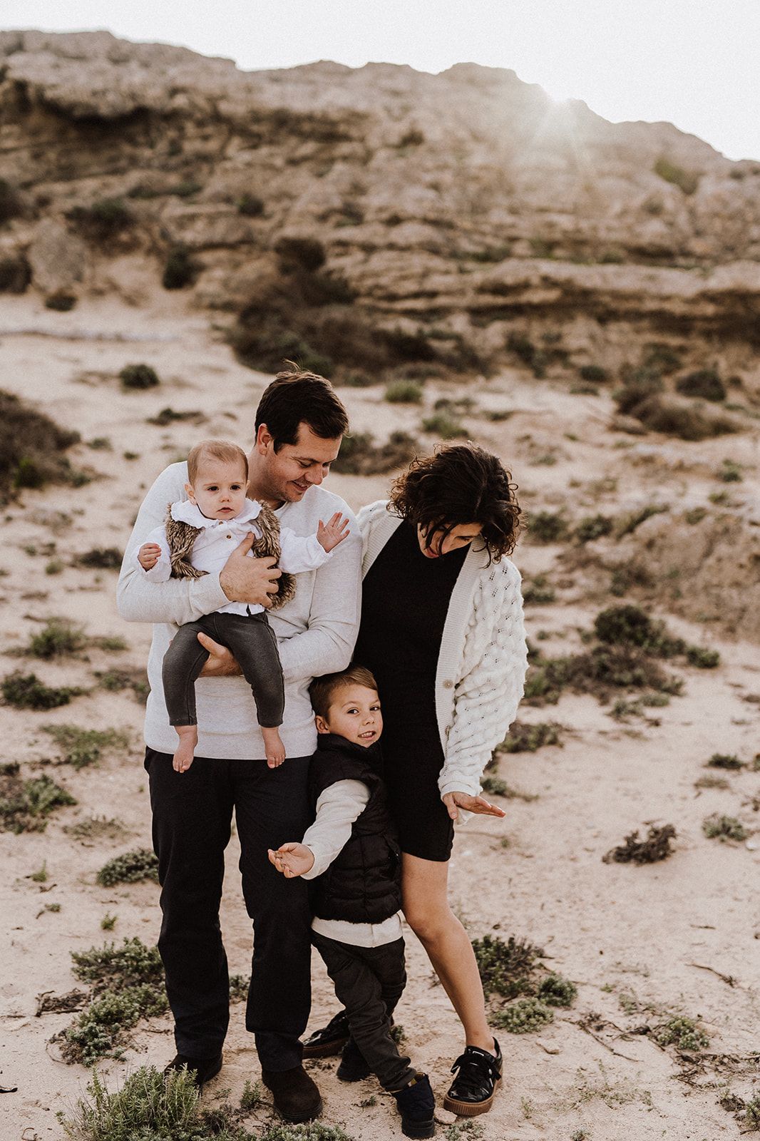 Family of four poses outdoors in desert landscape, parents holding young children with rocky cliffs and sunlight behind them.