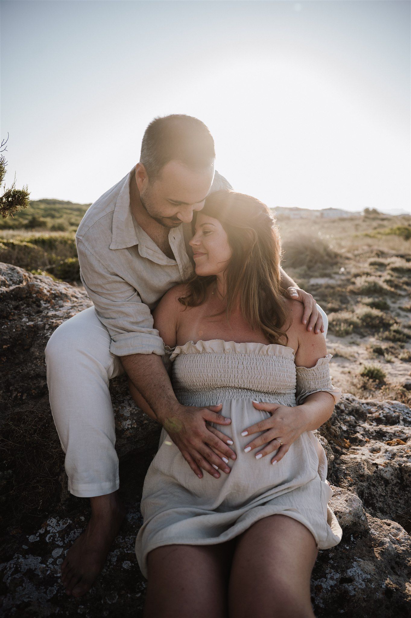 A couple shares a tender moment outdoors, the woman visibly pregnant, bathed in golden sunlight.