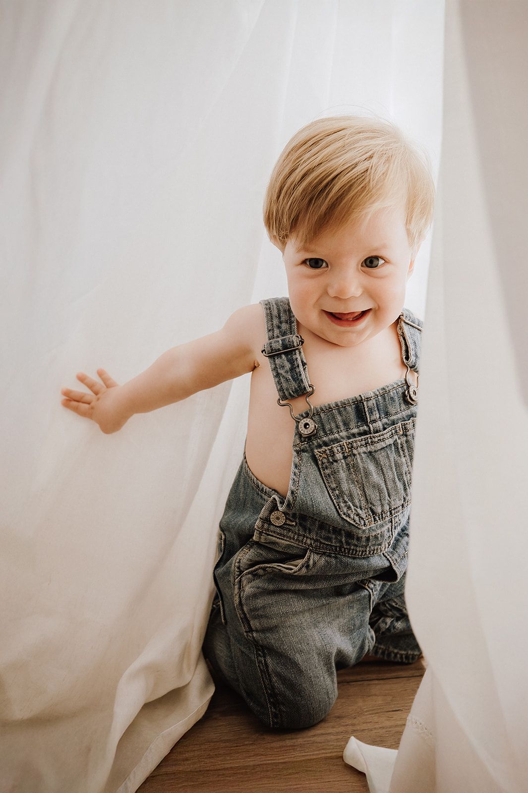 Smiling toddler boy in denim overalls sitting on wooden box surrounded by white sheer curtains.