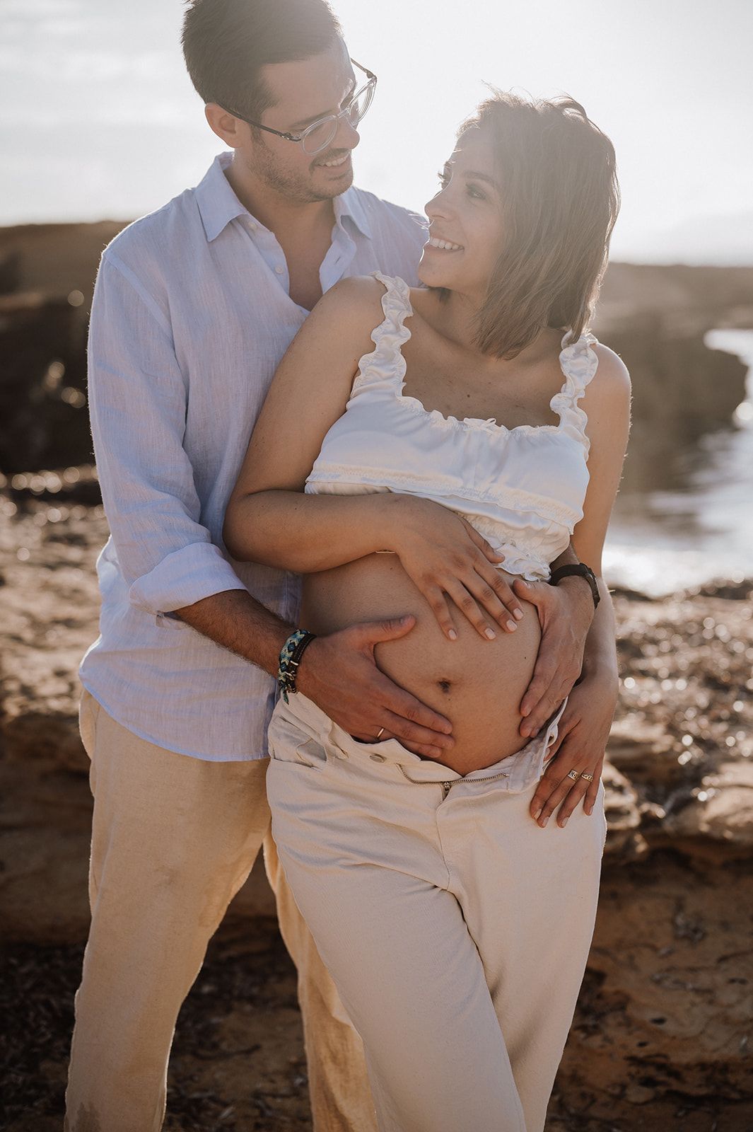Smiling couple embracing outdoors at sunset, hands cradling pregnant belly near rocky coastal backdrop.