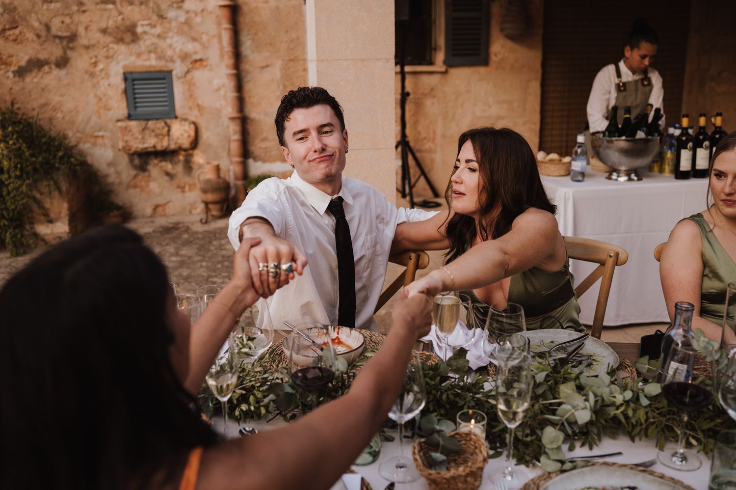 Wedding guests laughing and sharing food at an outdoor reception table with greenery centerpieces.