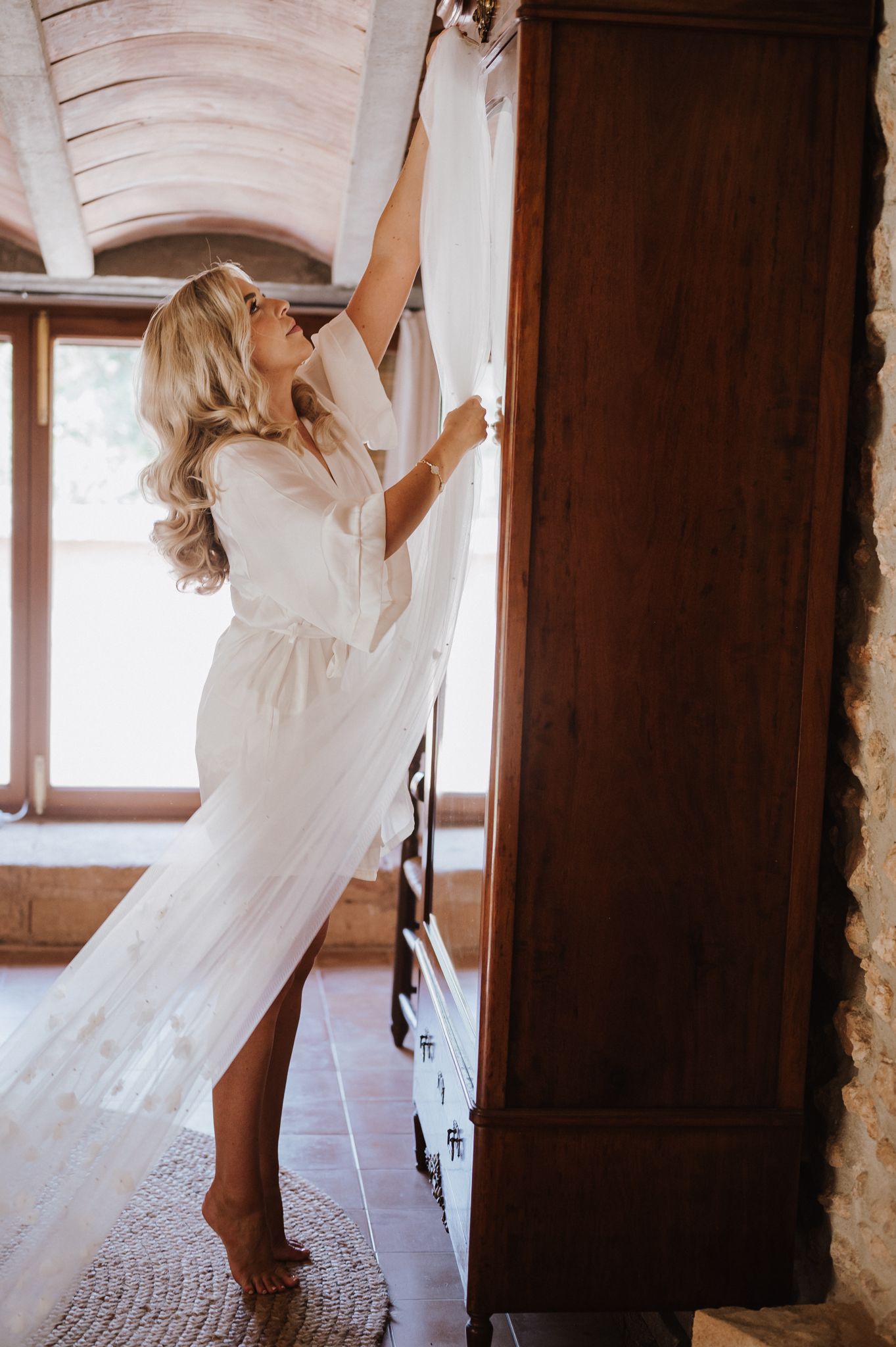 Blonde bride in a white robe reaching up to retrieve her wedding dress from a wooden armoire in a rustic room.