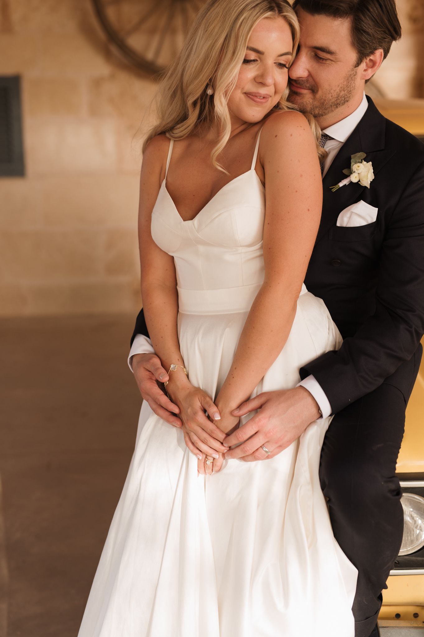 Groom embraces bride from behind in romantic portrait inside rustic venue, both wearing wedding attire.
