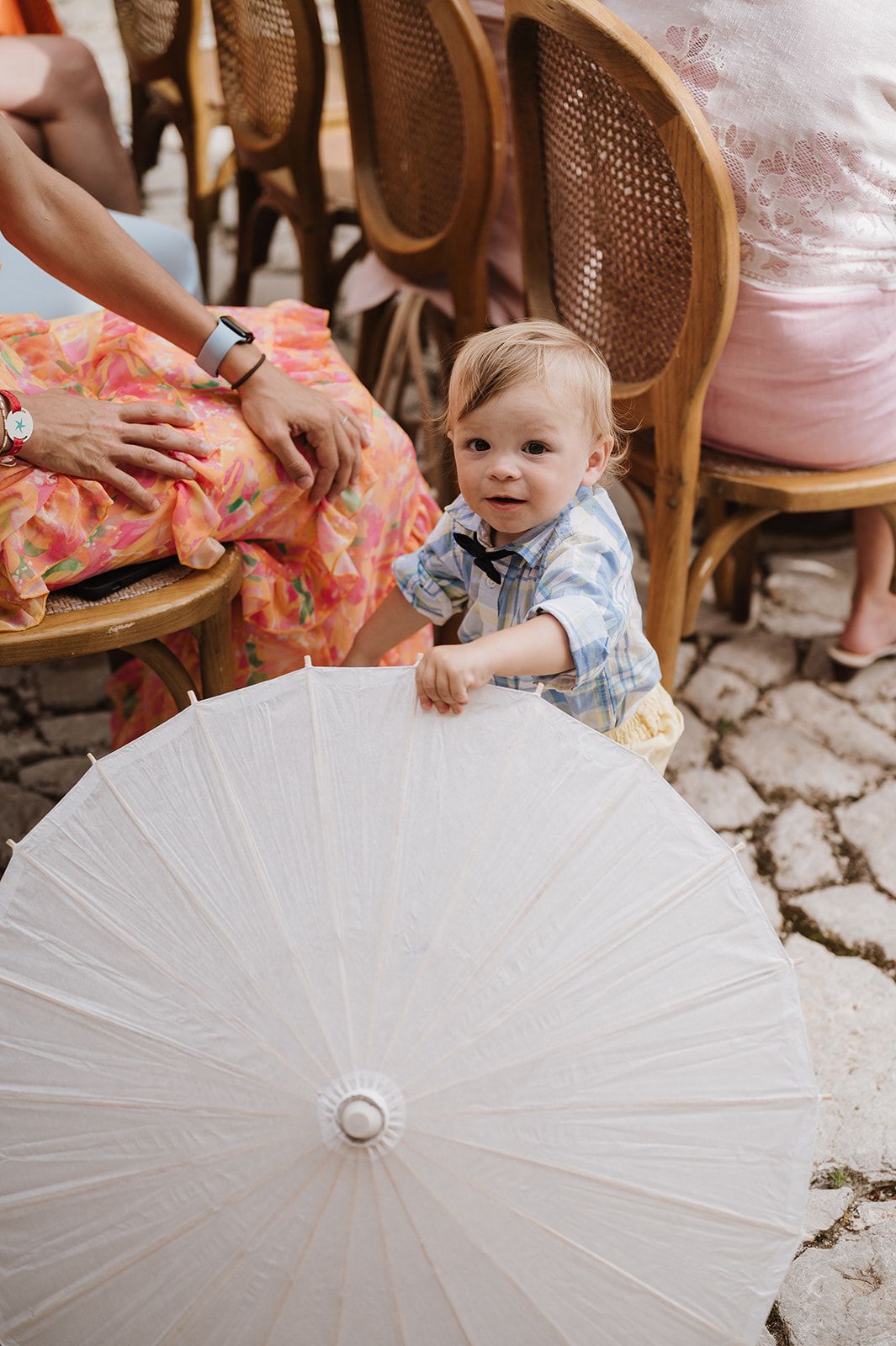 Adorable toddler in blue outfit holding a large white paper parasol at an outdoor wedding reception.