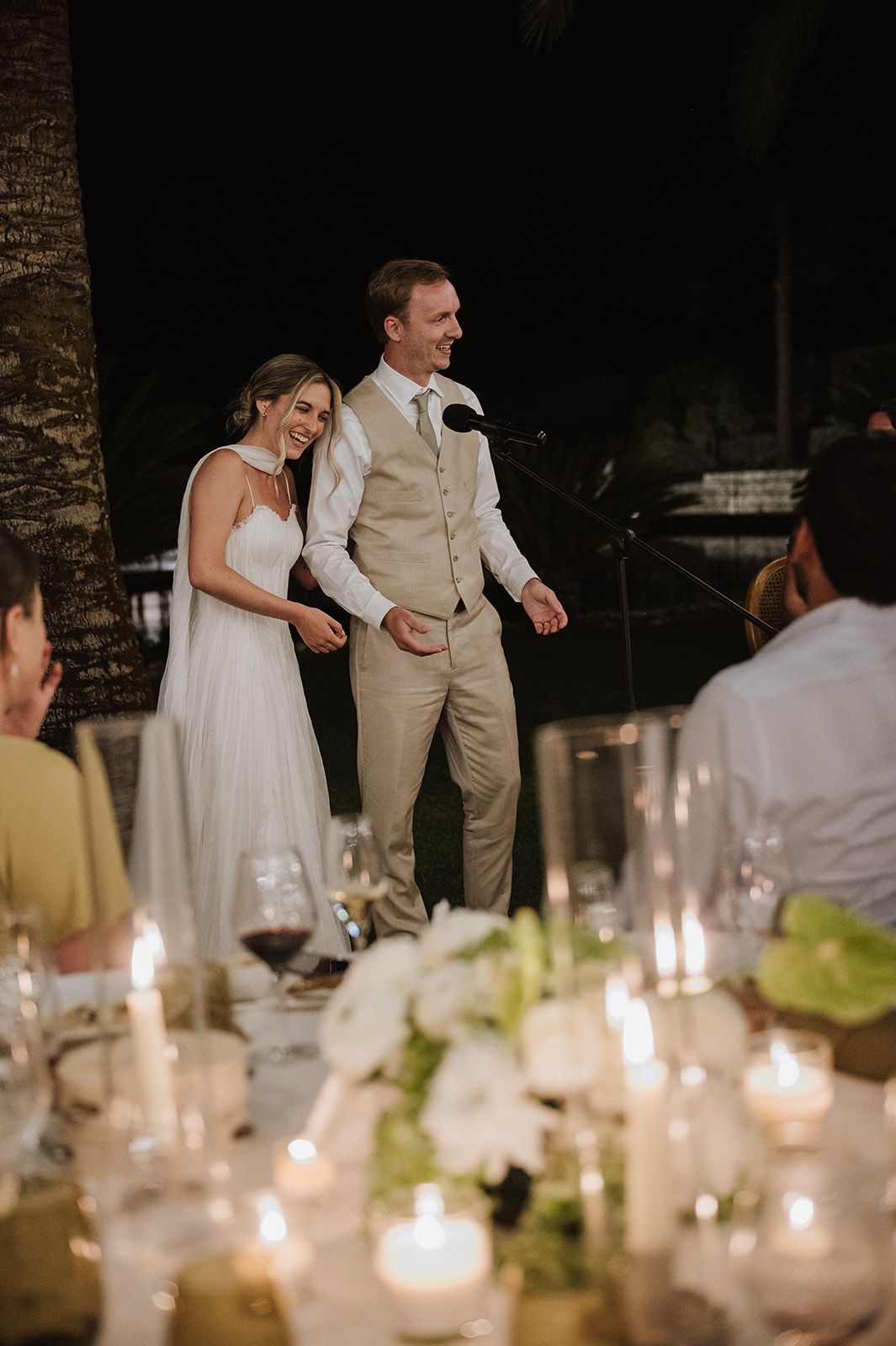 Bride and groom smile warmly at guests during candlelit outdoor evening wedding reception.