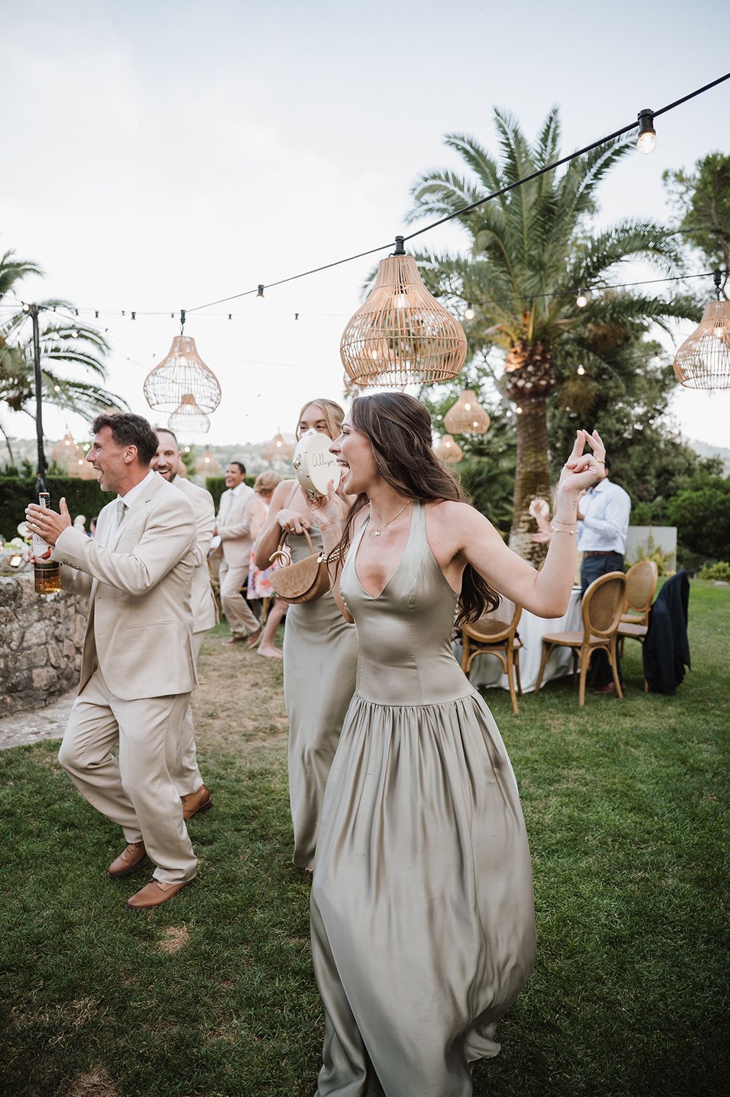 Bridesmaid in sage green satin gown dances joyfully under warm rattan pendant lights at outdoor wedding reception.