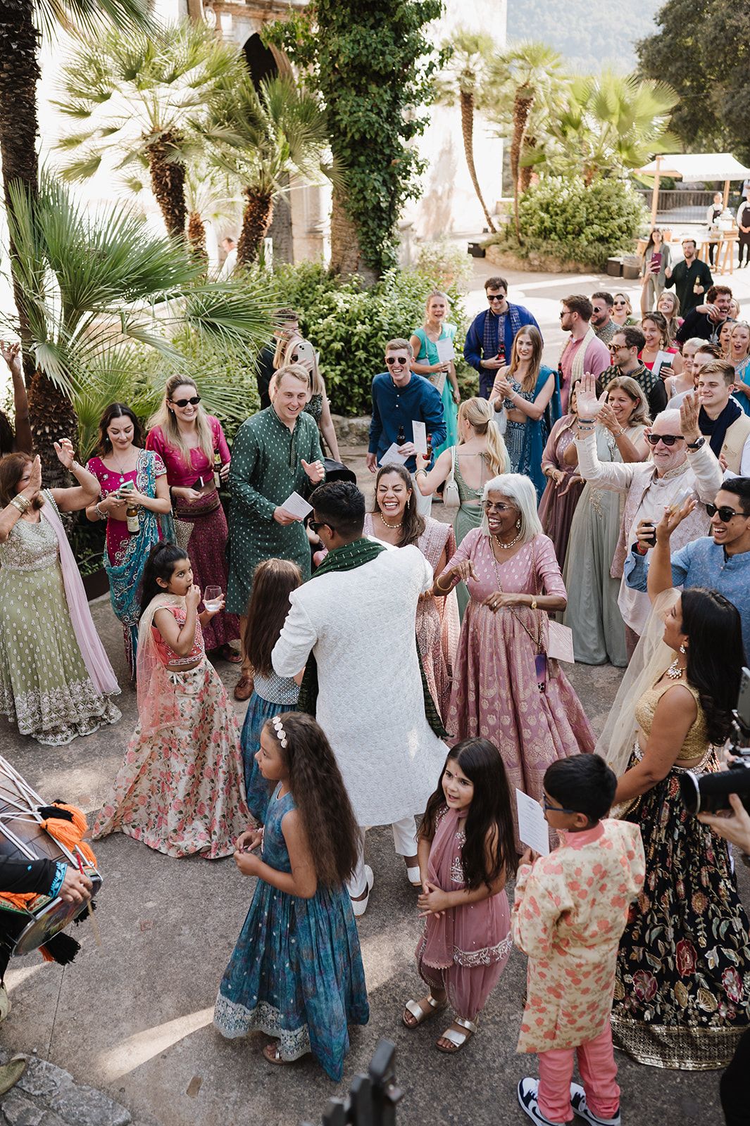 Wedding guests dancing joyfully in circle during Indian baraat ceremony in garden setting.