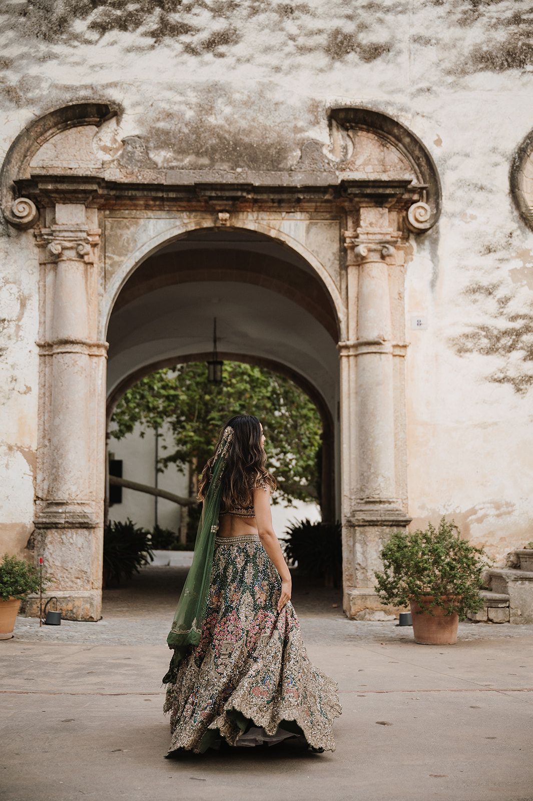 Bride in ornate green floral lehenga twirling before a stone archway at an elegant wedding venue.