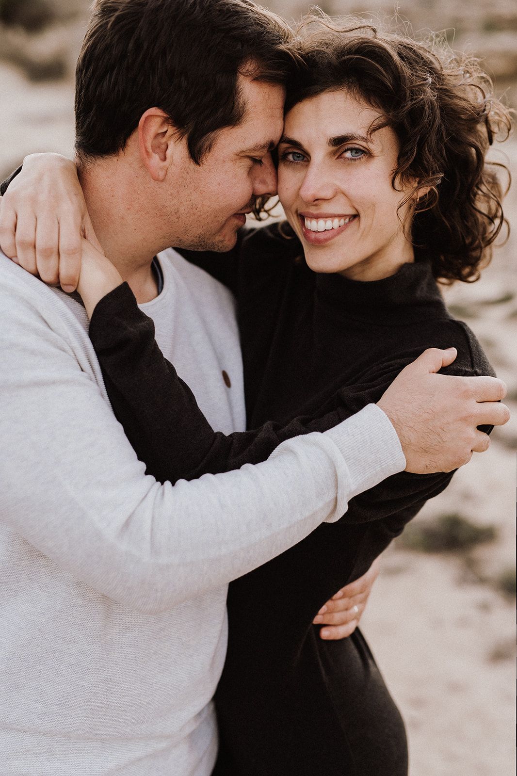 A smiling couple embraces outdoors, woman in black turtleneck with curly hair, man in white sweatshirt nuzzling her cheek.