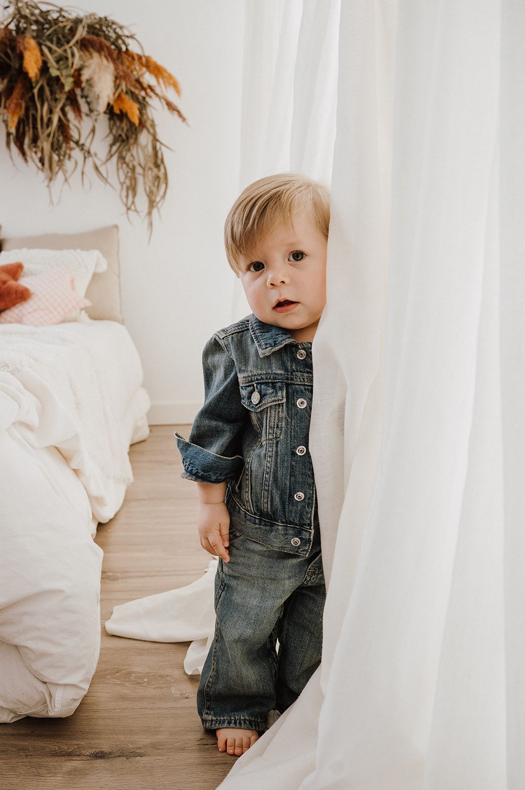 Curious toddler boy in denim jacket and jeans peeking around a white curtain in a boho bedroom.