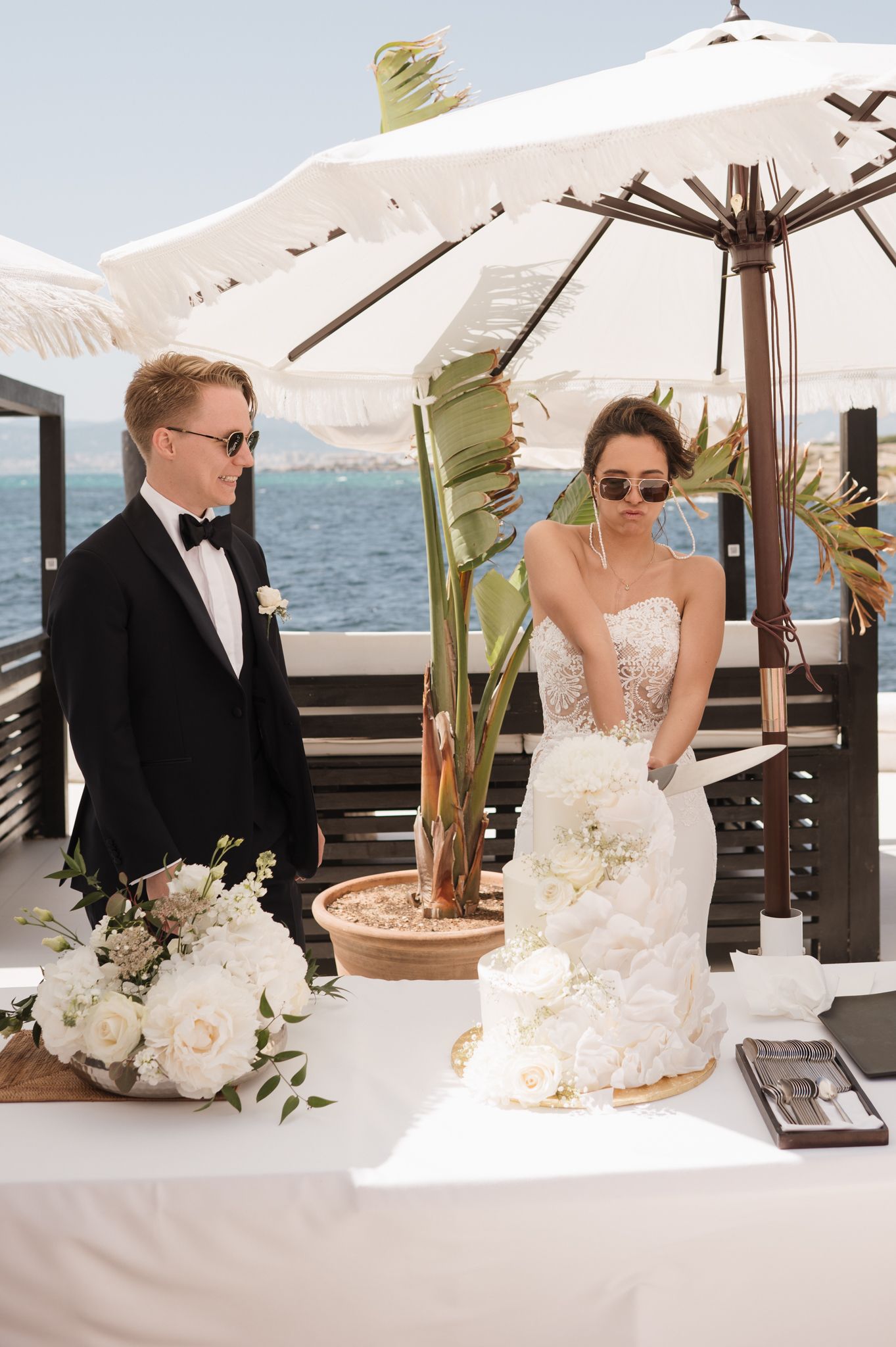 Bride and groom in sunglasses cut white wedding cake at oceanside reception.