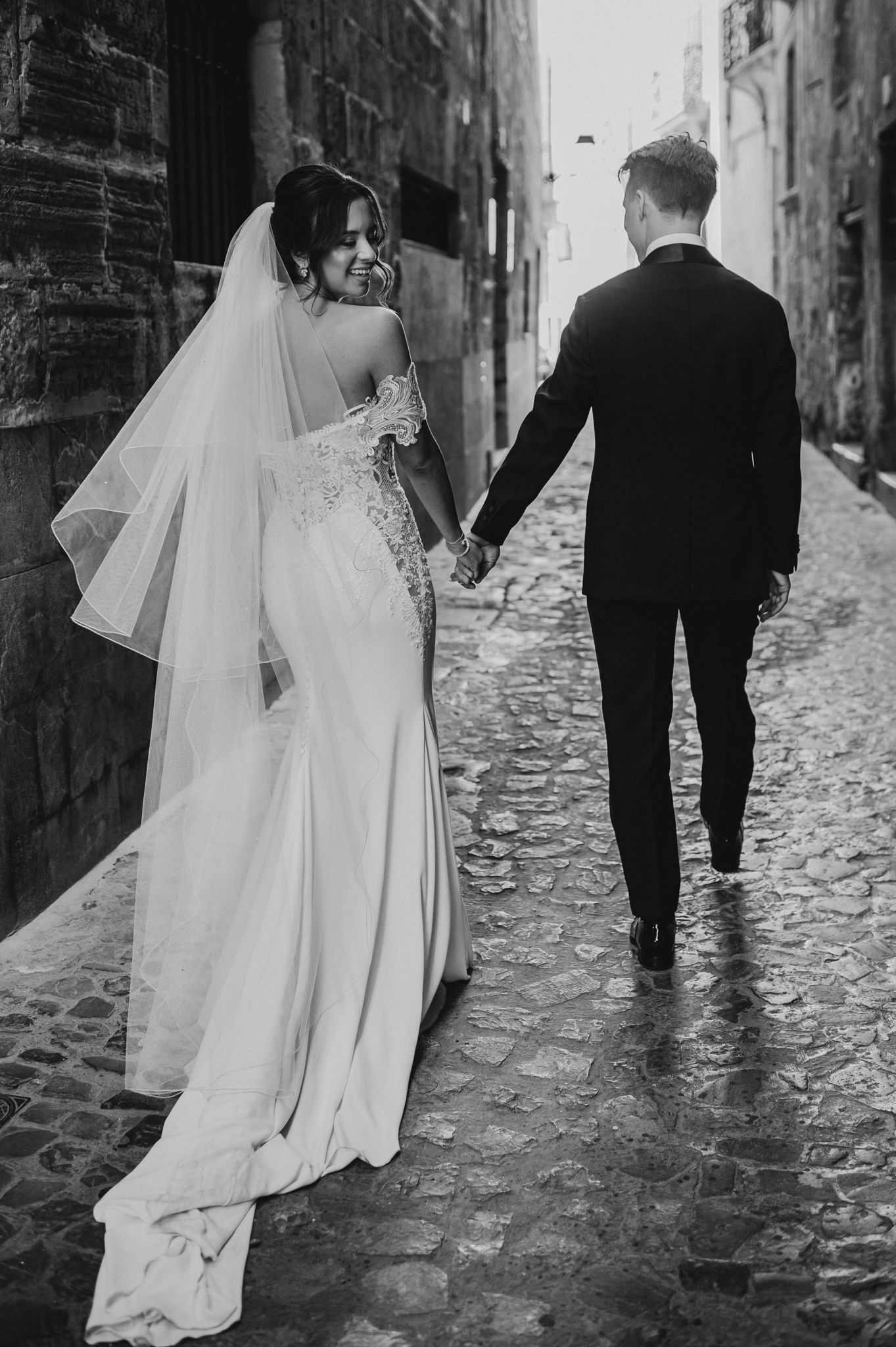 Bride and groom walk hand in hand down a cobblestone alley in a romantic black and white wedding photo.