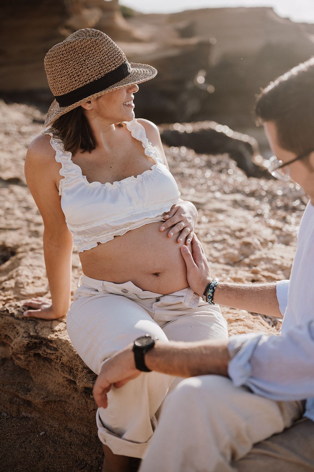 Pregnant woman in white crop top and hat sitting on rocks while partner touches her belly.