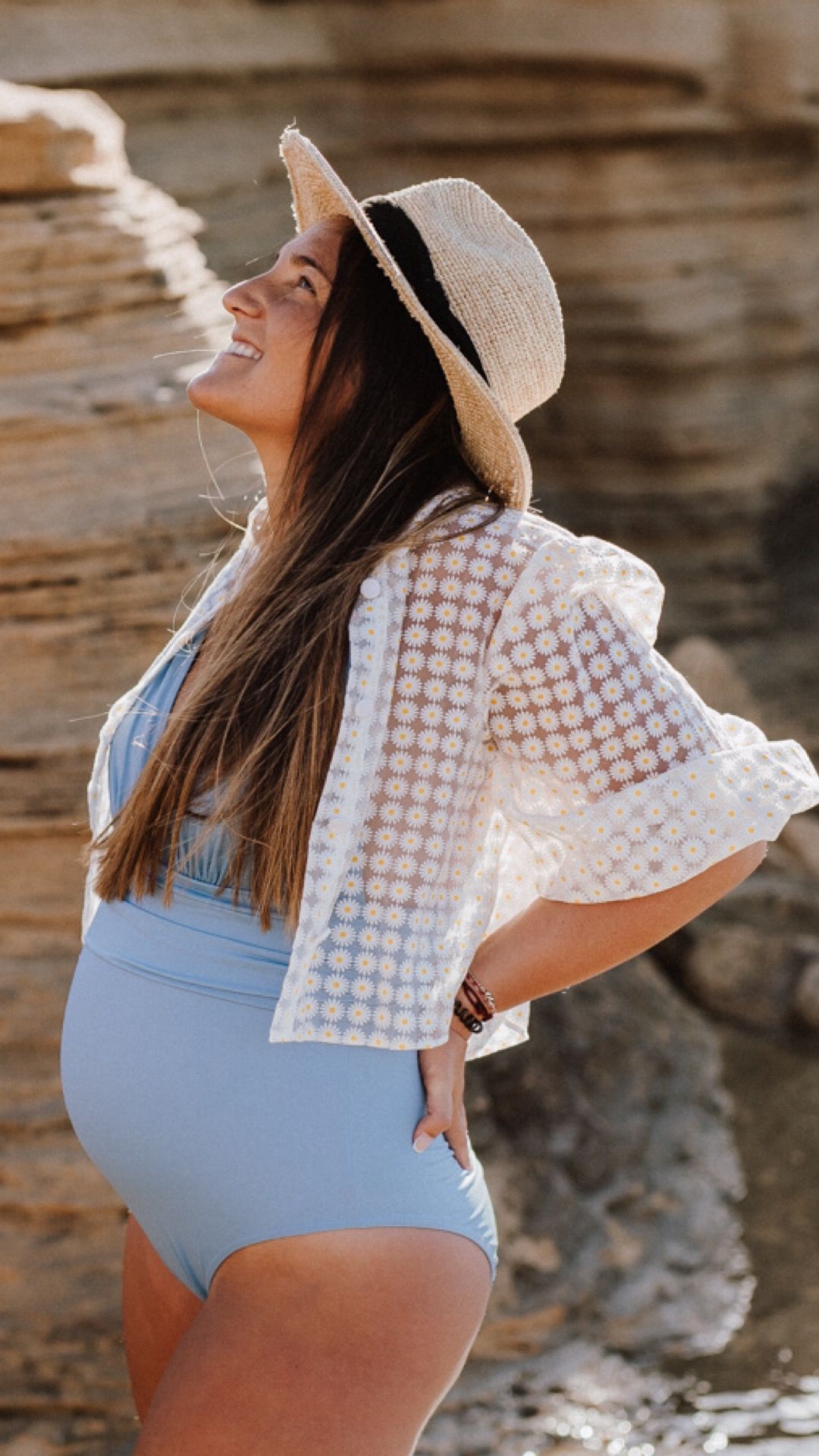 Smiling pregnant woman in light blue swimsuit, lace cover-up, and straw hat posing by rocky cliffs.
