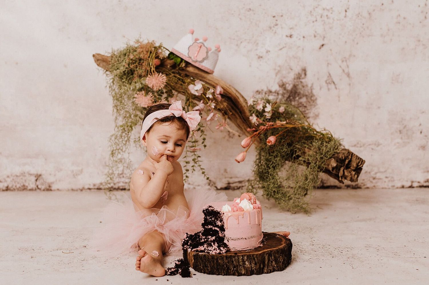 Baby girl in pink tutu smashing chocolate birthday cake, seated on wood slice with floral branch backdrop.