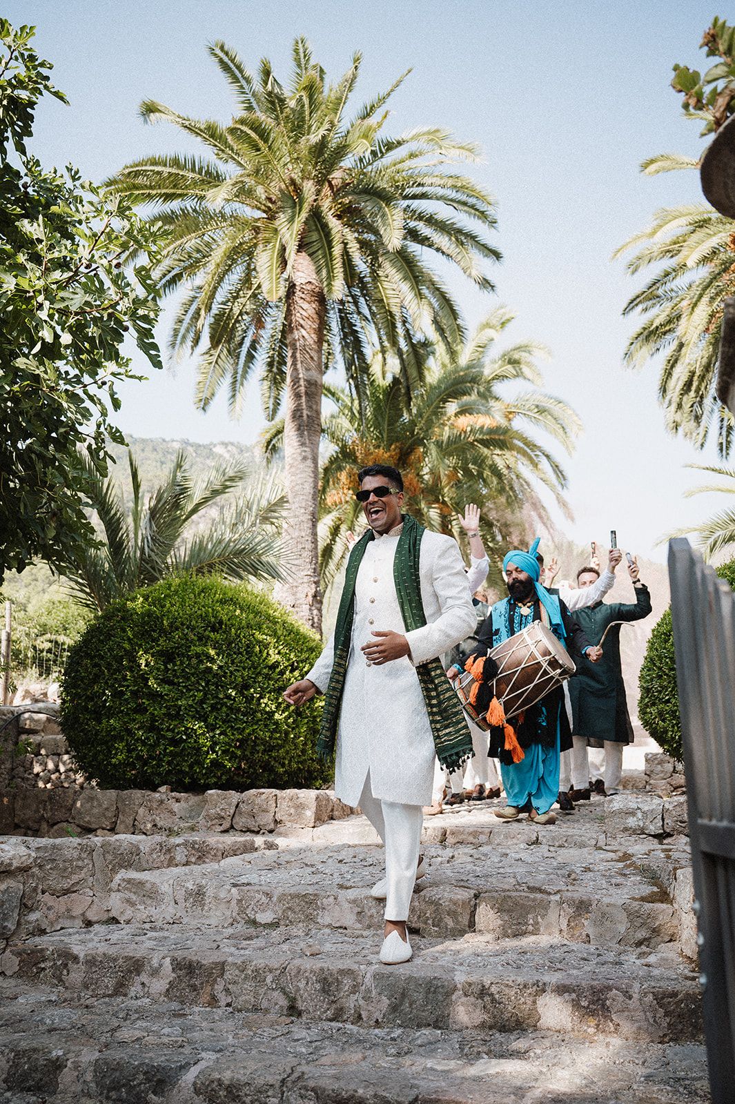 Groom in white sherwani leading baraat procession with dhol players on stone steps amid palm trees.