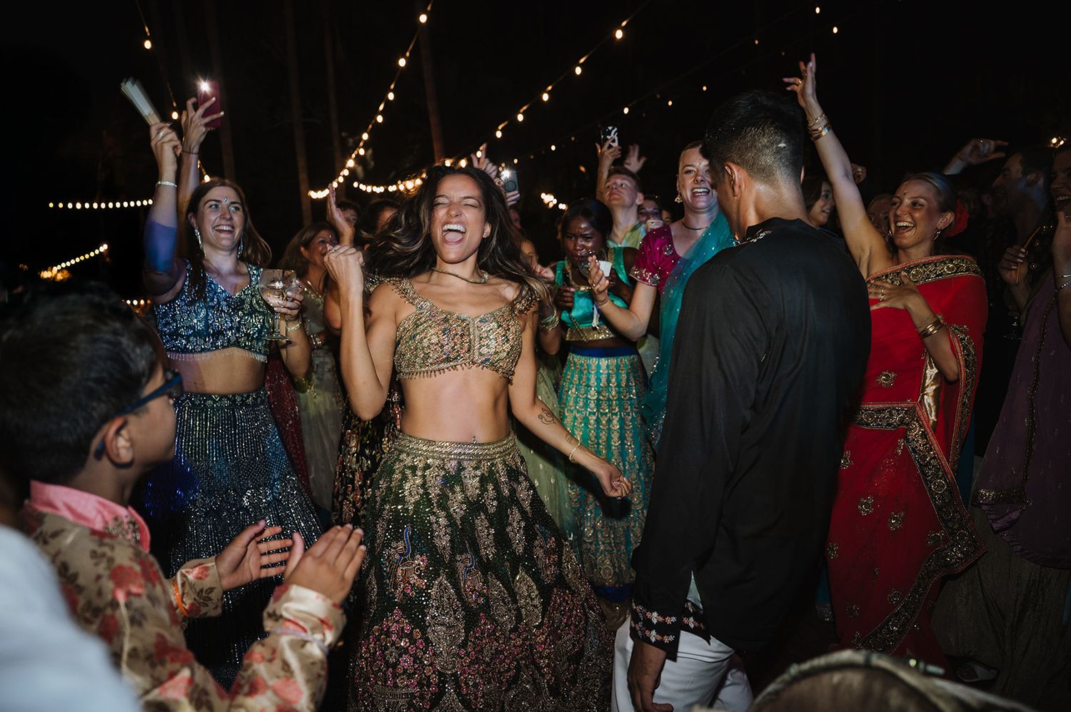 Woman in green and gold lehenga laughs and dances joyfully at an outdoor Indian wedding reception.