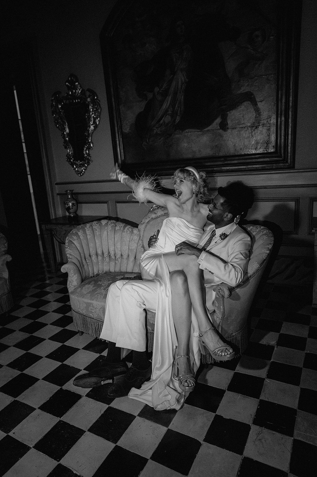 Black and white photo of a bride and groom sitting on a vintage sofa in an elegant room with checkered floors.