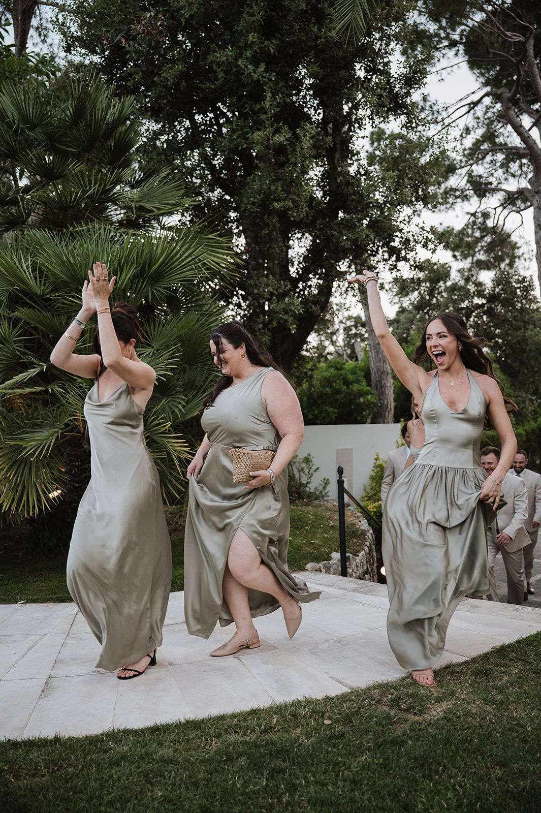 Three bridesmaids in sage green satin dresses dancing joyfully outdoors at a garden wedding reception.