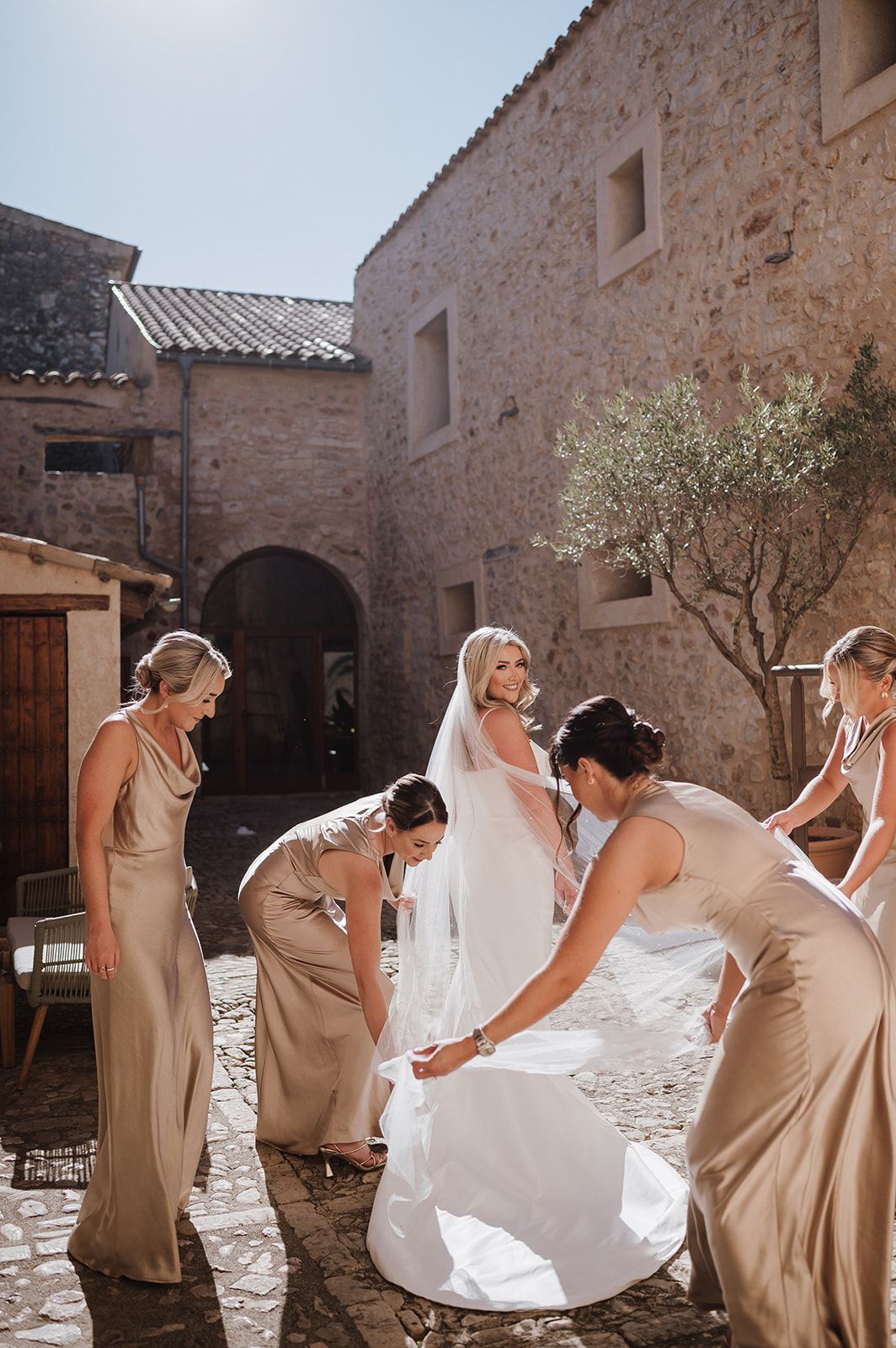 Bridesmaids in gold satin gowns arrange a bride's white wedding dress train in a rustic stone courtyard.