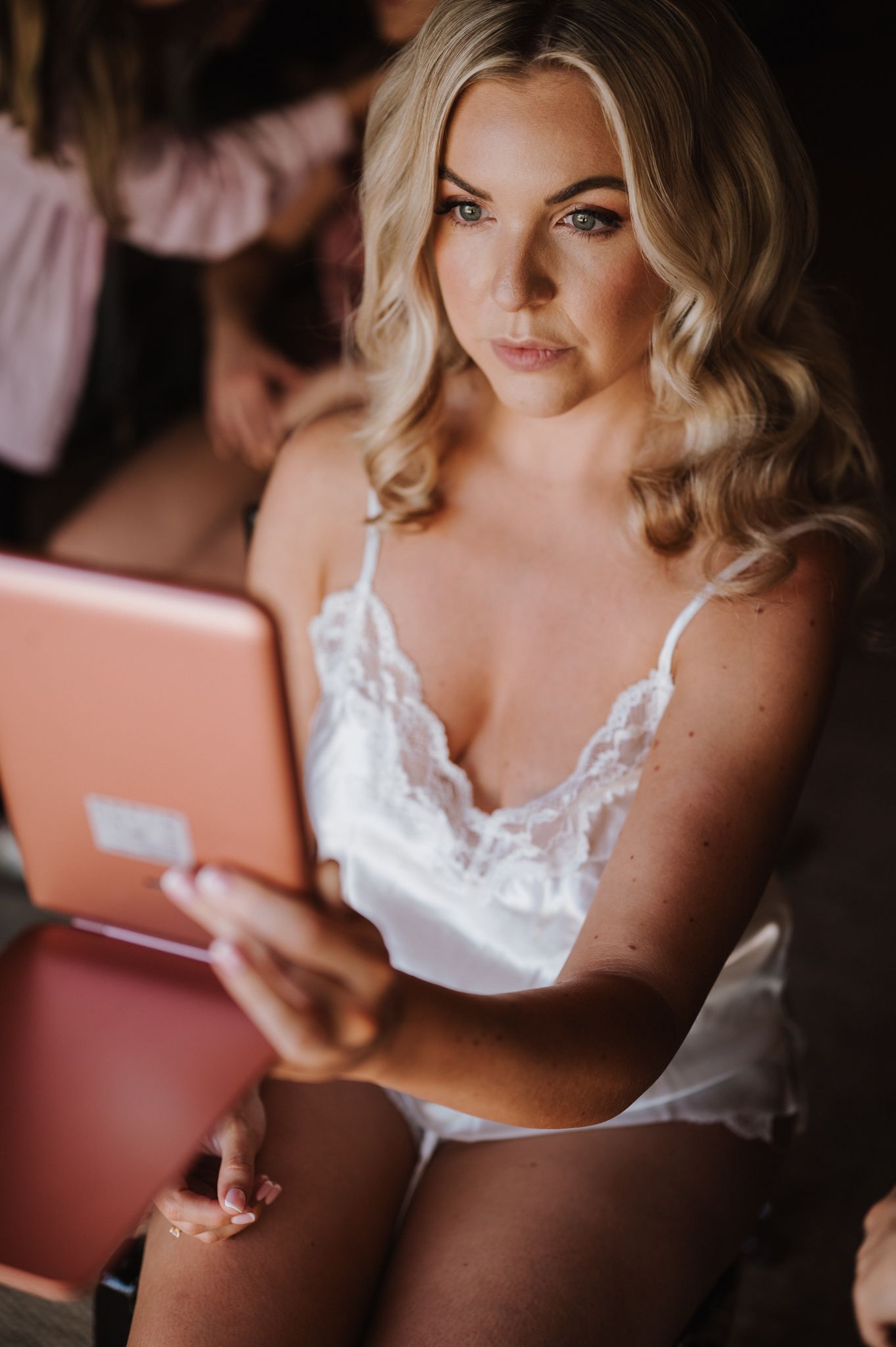 Blonde woman in white lace lingerie holding a pink tablet, sitting during bridal preparation getting-ready session.