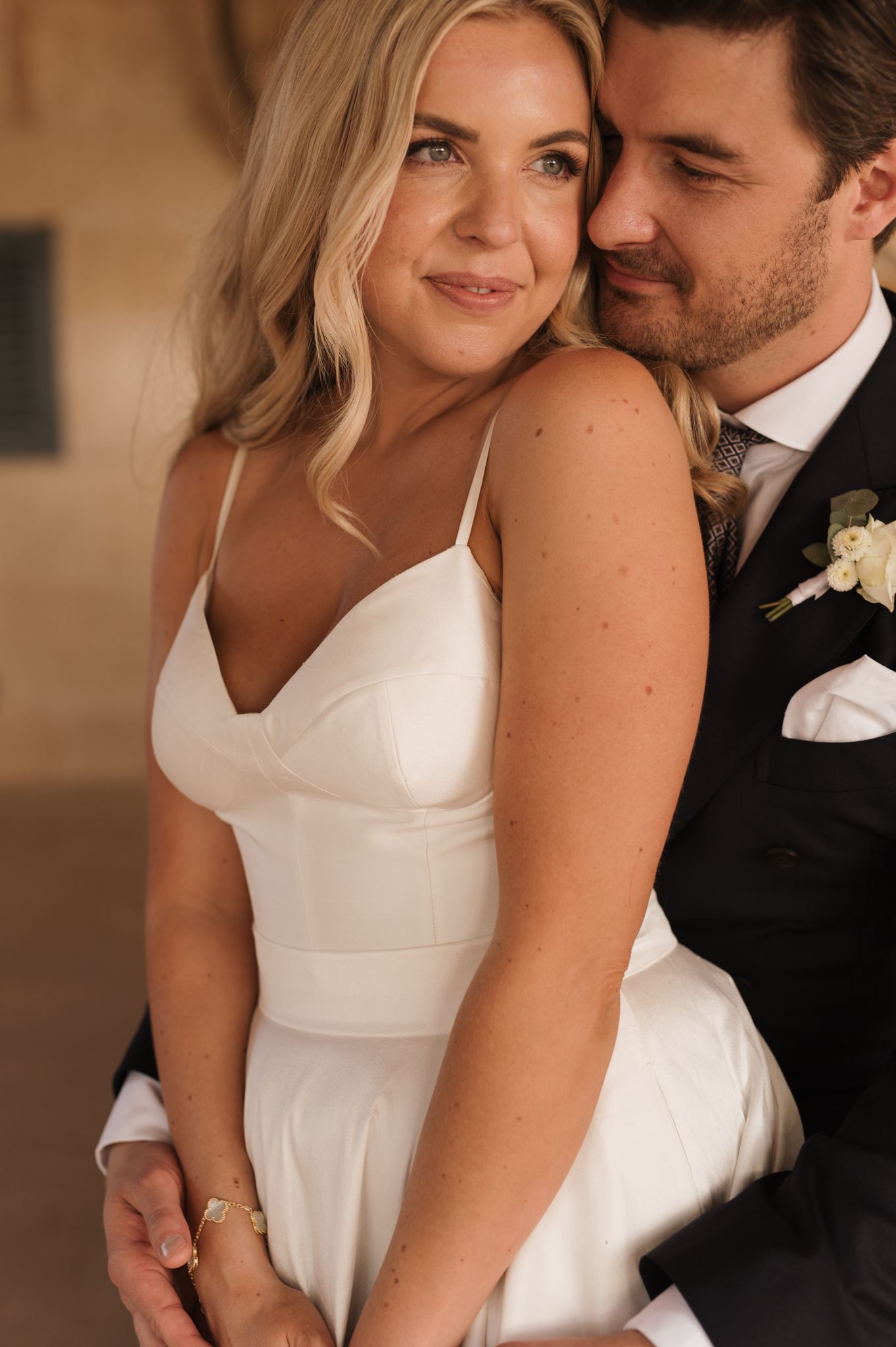 Bride in white spaghetti strap gown smiling as groom in dark suit embraces her from behind.