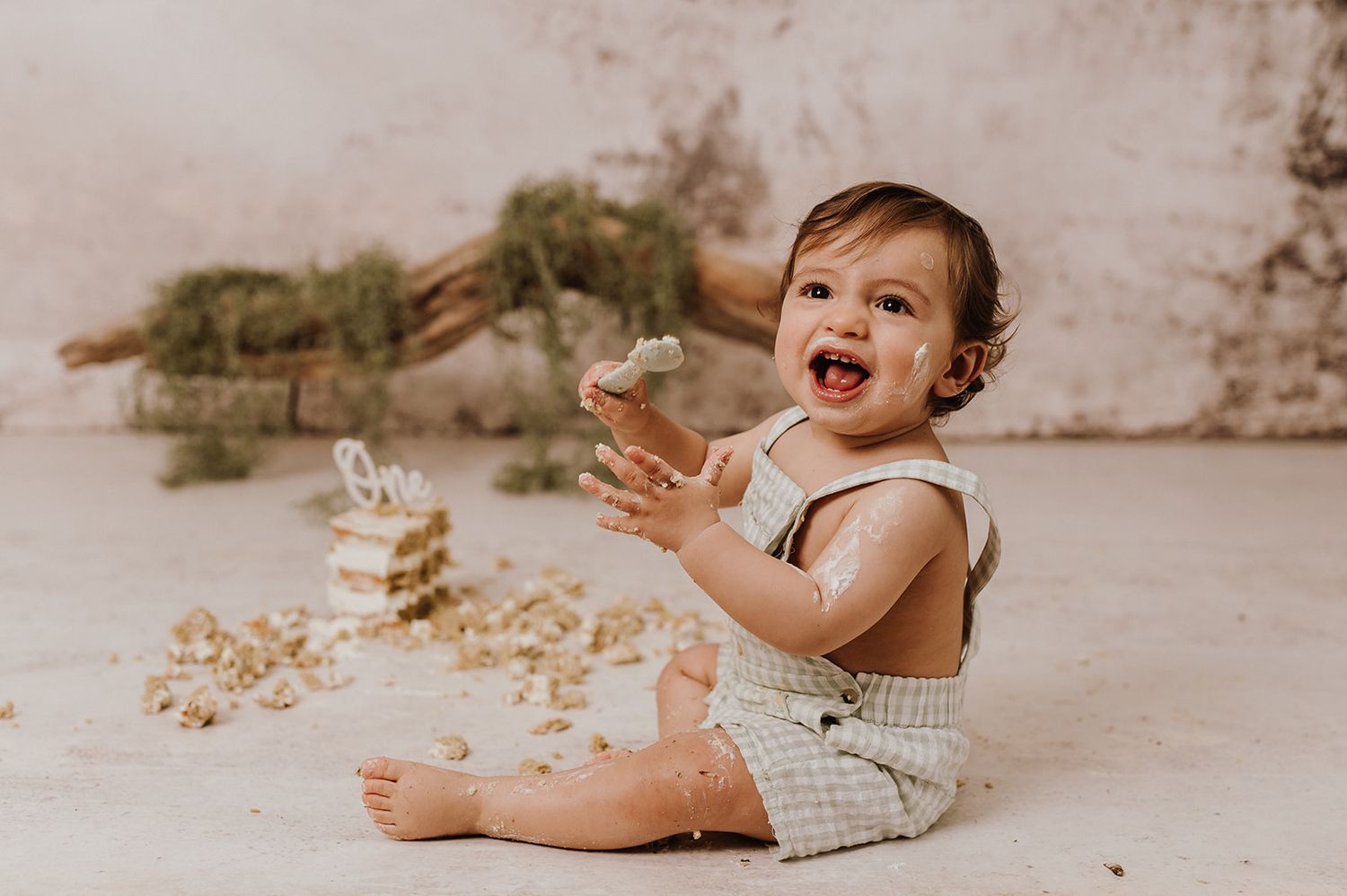A joyful baby in a checkered romper laughs while smashing a cake, sitting on a white floor.