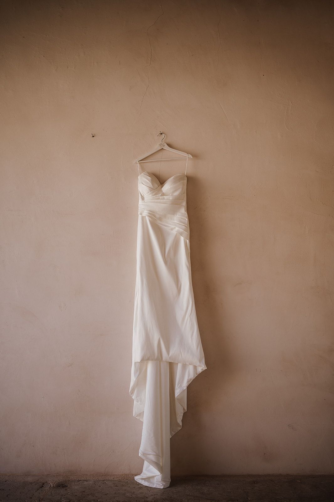 Elegant strapless ivory wedding gown hanging on hanger against beige wall.
