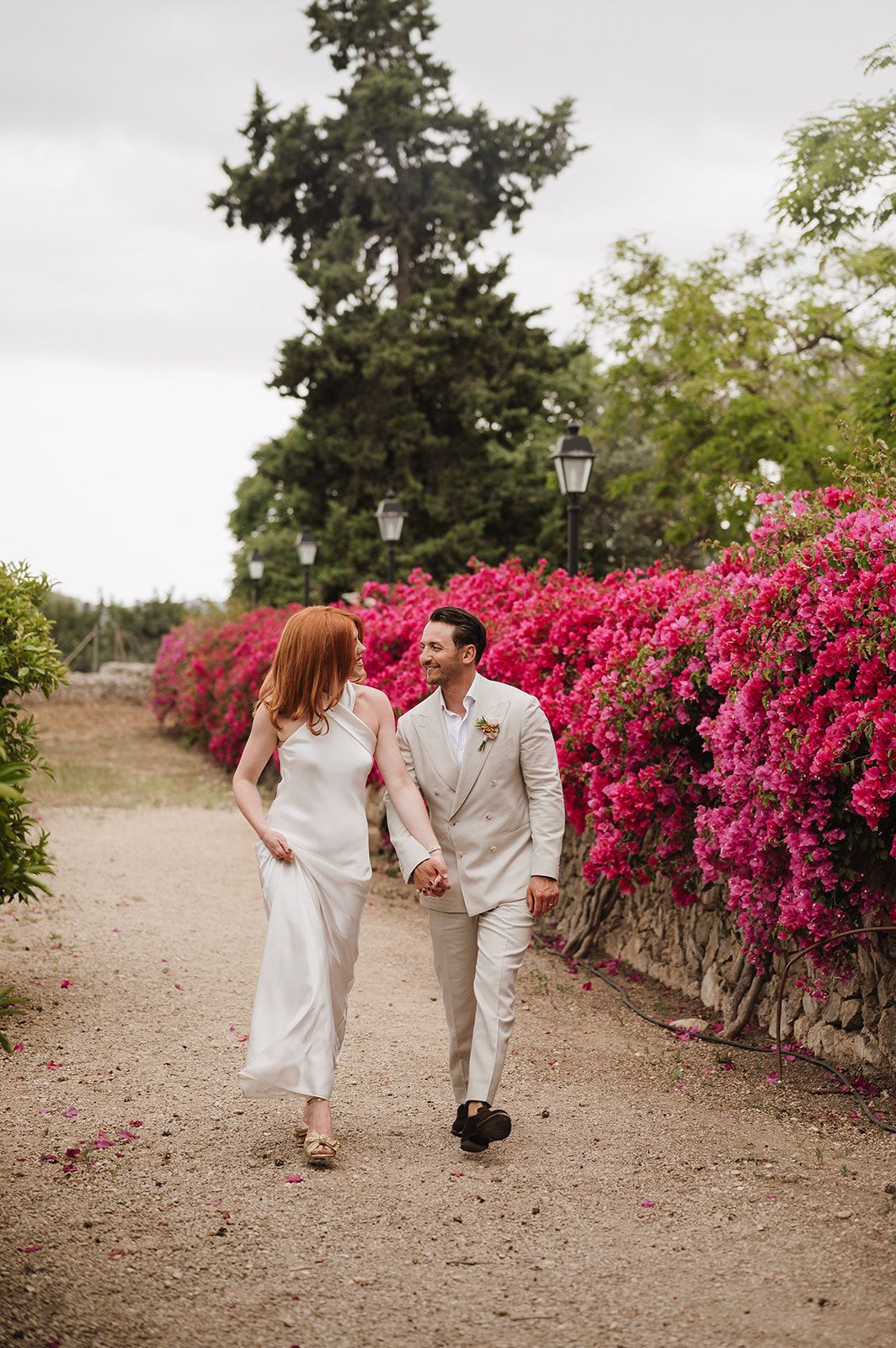 Bride and groom holding hands walking along a gravel path lined with vivid pink bougainvillea blooms.
