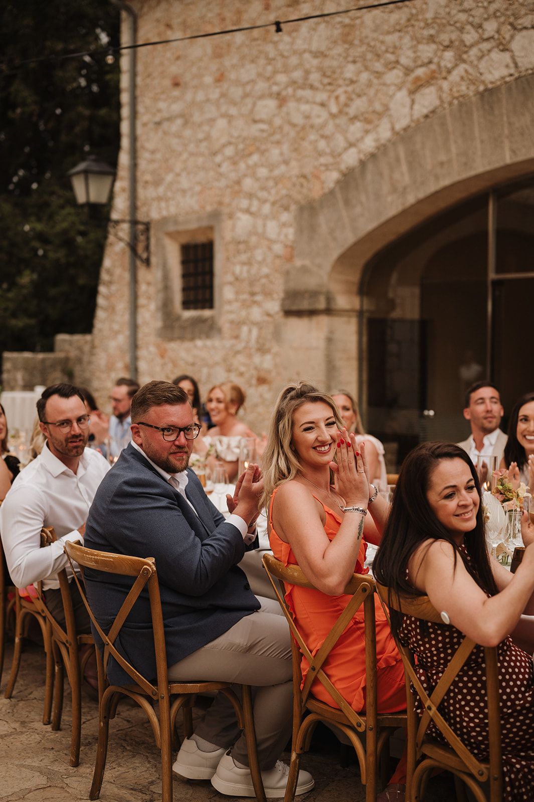 Wedding guests applaud and smile at outdoor rustic stone courtyard reception dinner celebration.