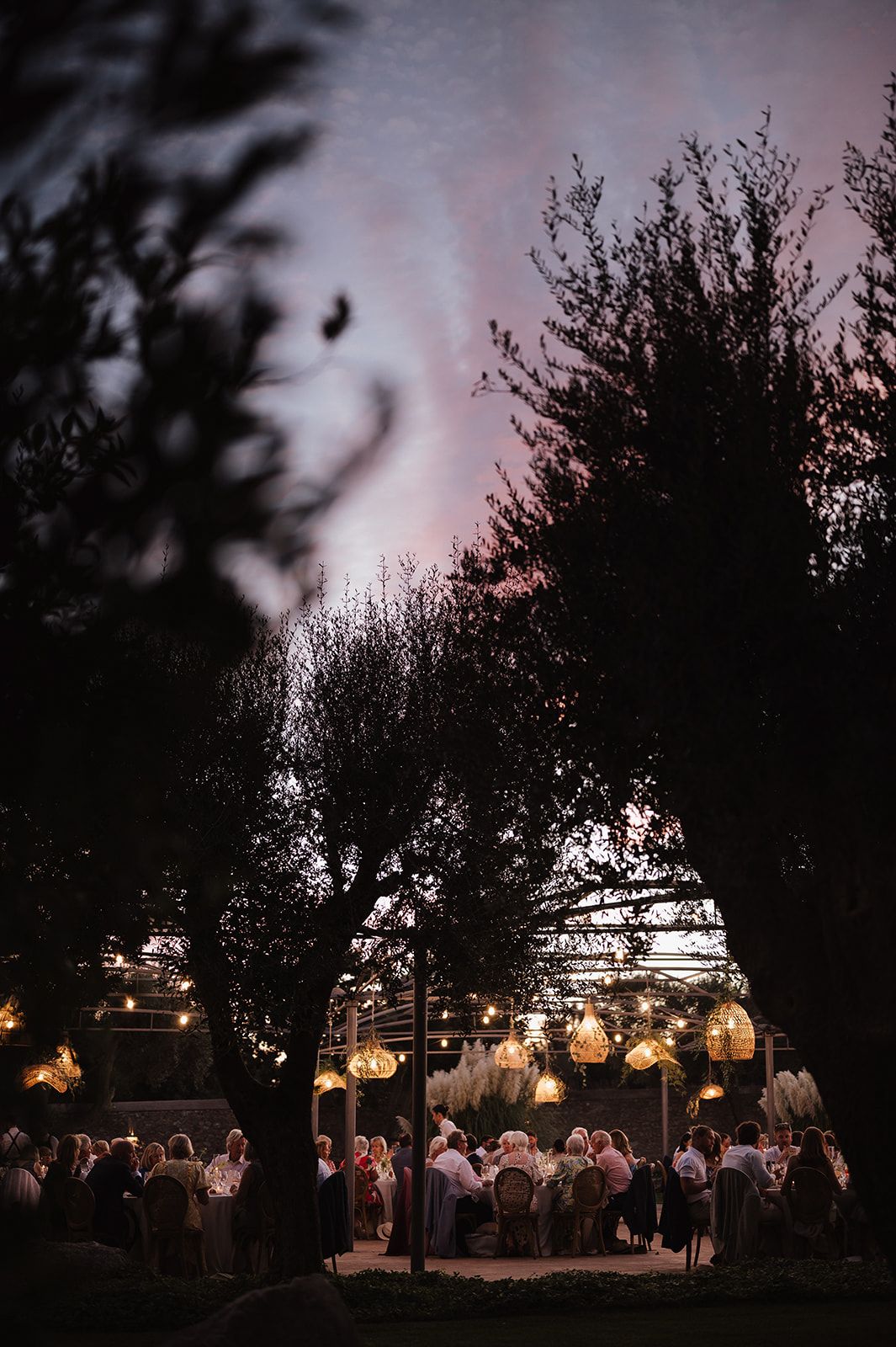 Guests gather under warm glowing lanterns at an elegant outdoor wedding reception at dusk.