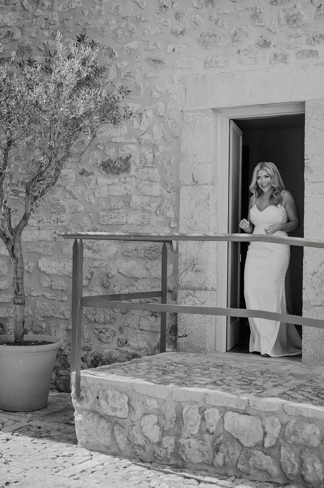 Bride in white gown standing in stone building doorway with olive tree beside rustic steps.