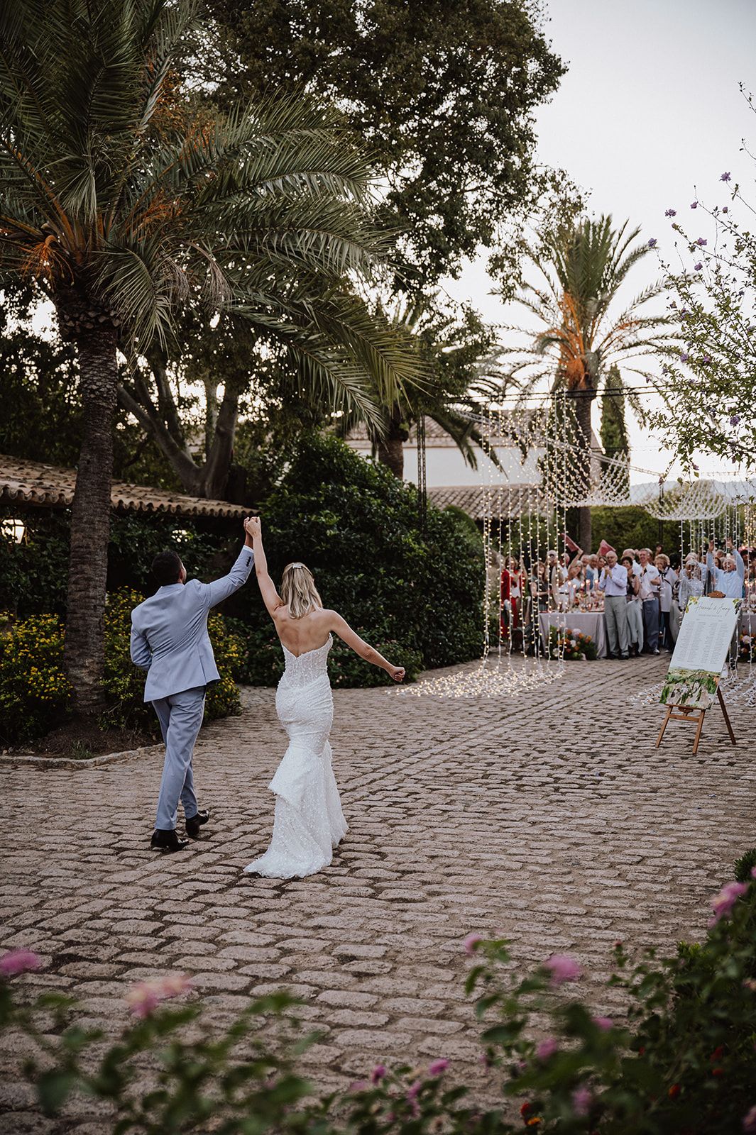 Bride and groom hold hands on cobblestone path at outdoor wedding reception with guests and palm trees.