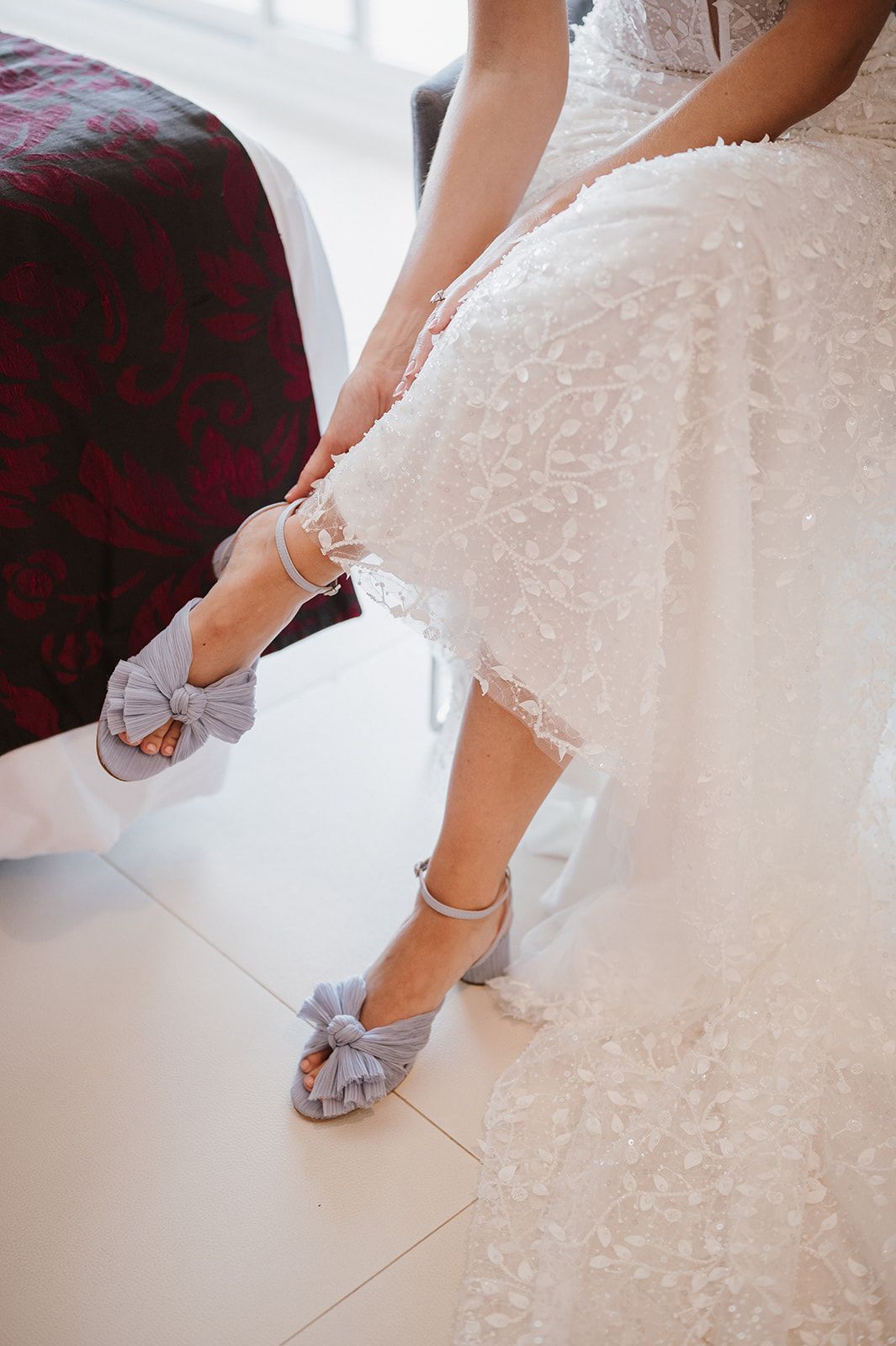 Bride being helped into blue bow-detail block heel wedding shoes with lace gown detail.