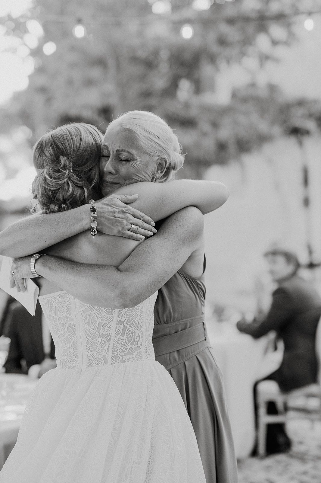 Bride in lace wedding gown shares an emotional hug with an older woman at the reception.