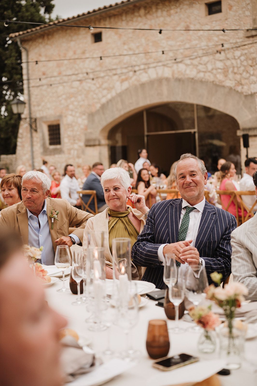 Wedding guests in elegant attire smile and laugh at an outdoor rustic stone venue reception dinner.