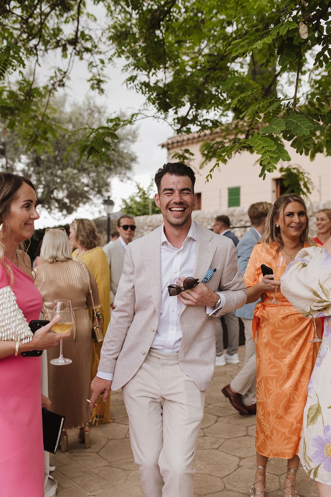 Smiling man in beige suit walks confidently through a crowd of elegantly dressed wedding guests outdoors.