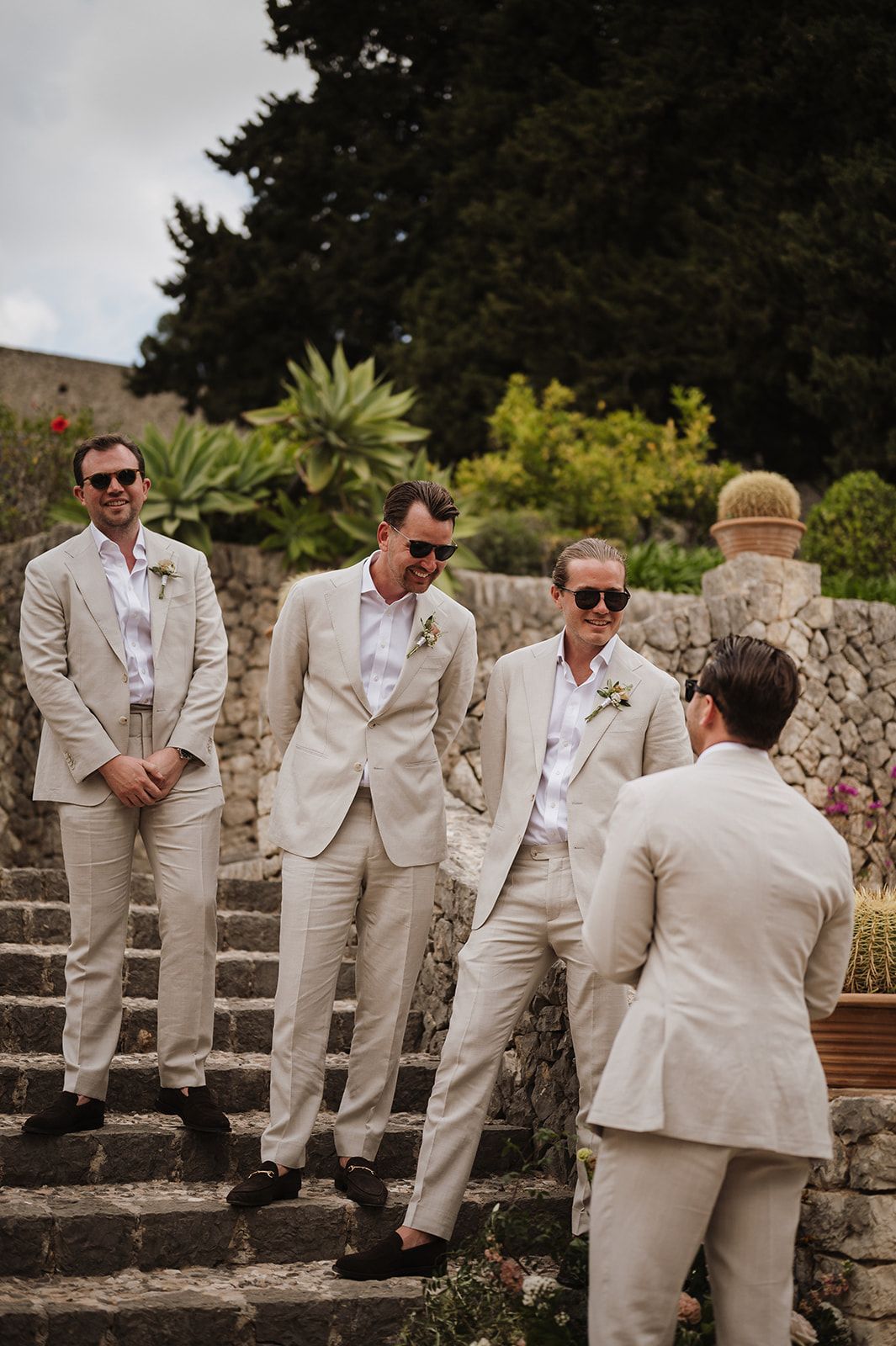 Three groomsmen in cream linen suits with boutonnieres pose on stone steps at outdoor wedding.