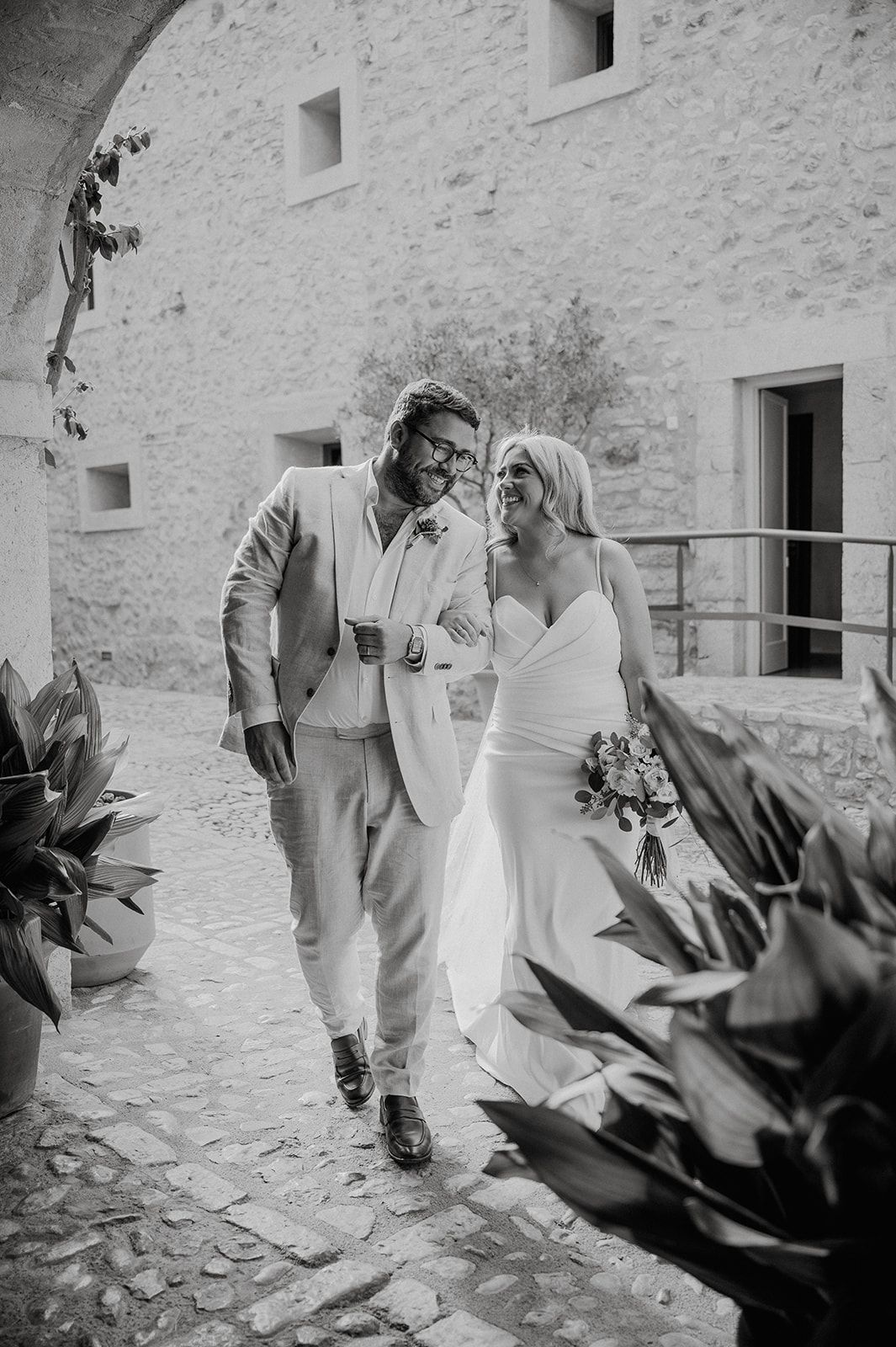 Bride and groom walking and smiling together through a rustic stone courtyard in black and white photo.
