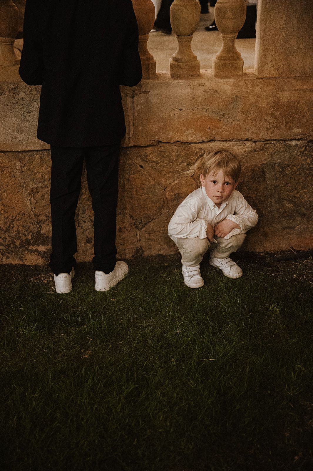 Adorable young child in white outfit crouches on grass beside adult in black suit at wedding venue.
