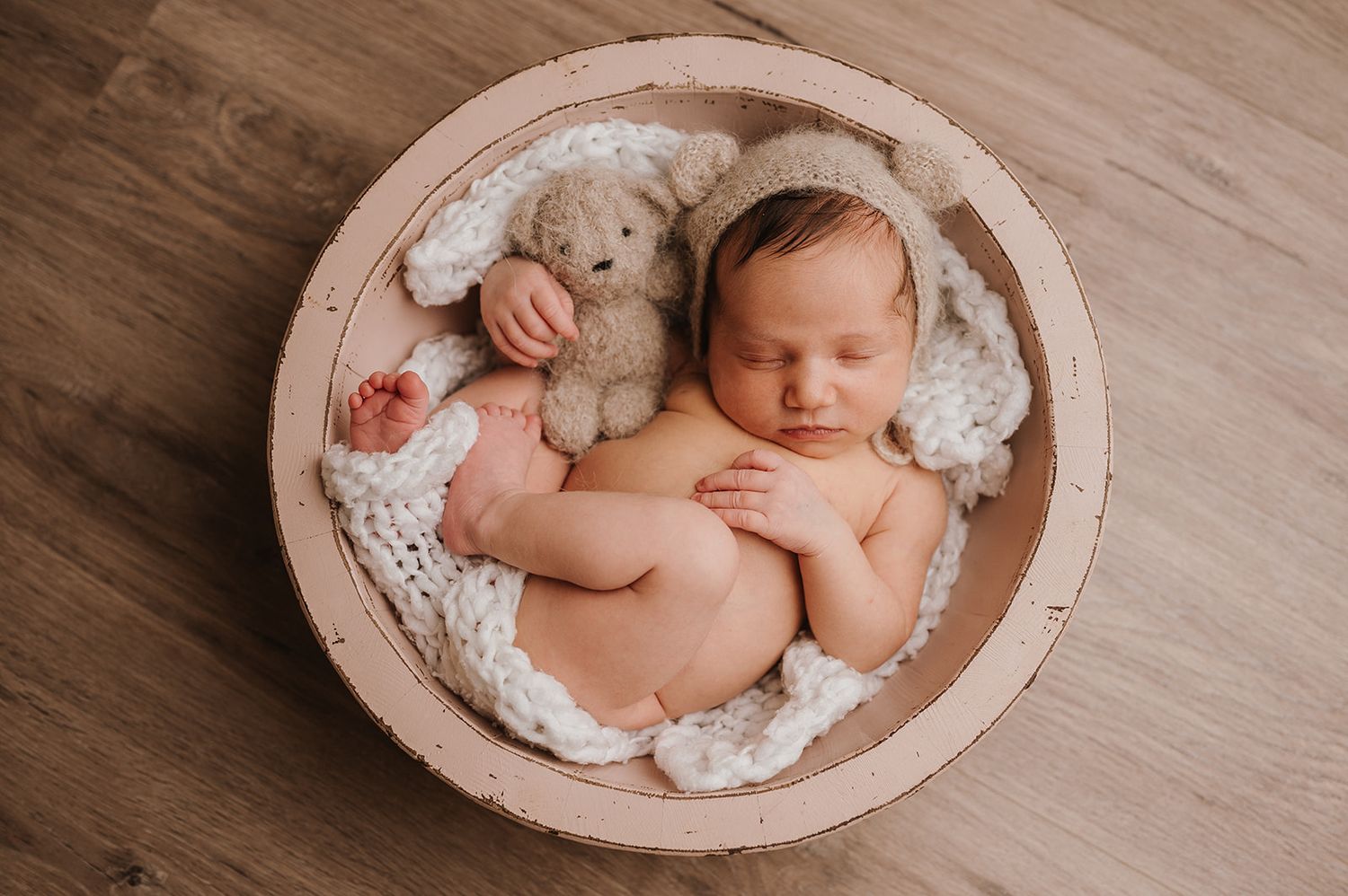 Newborn baby sleeping peacefully in a wooden bowl with a soft teddy bear on a white knit blanket.