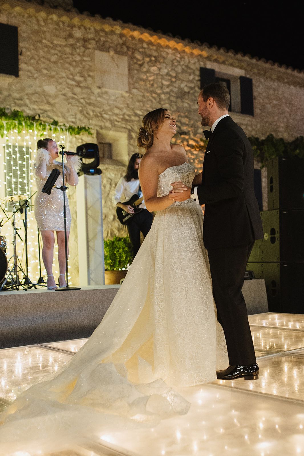 Bride and groom share their first dance outdoors at night, with a live band performing behind them at a romantic wedding reception.