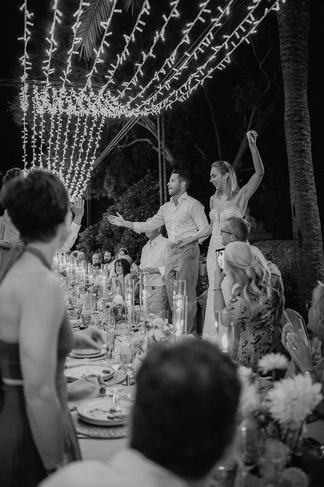 Bride and groom stand joyfully at their candlelit outdoor reception table under a canopy of fairy lights.
