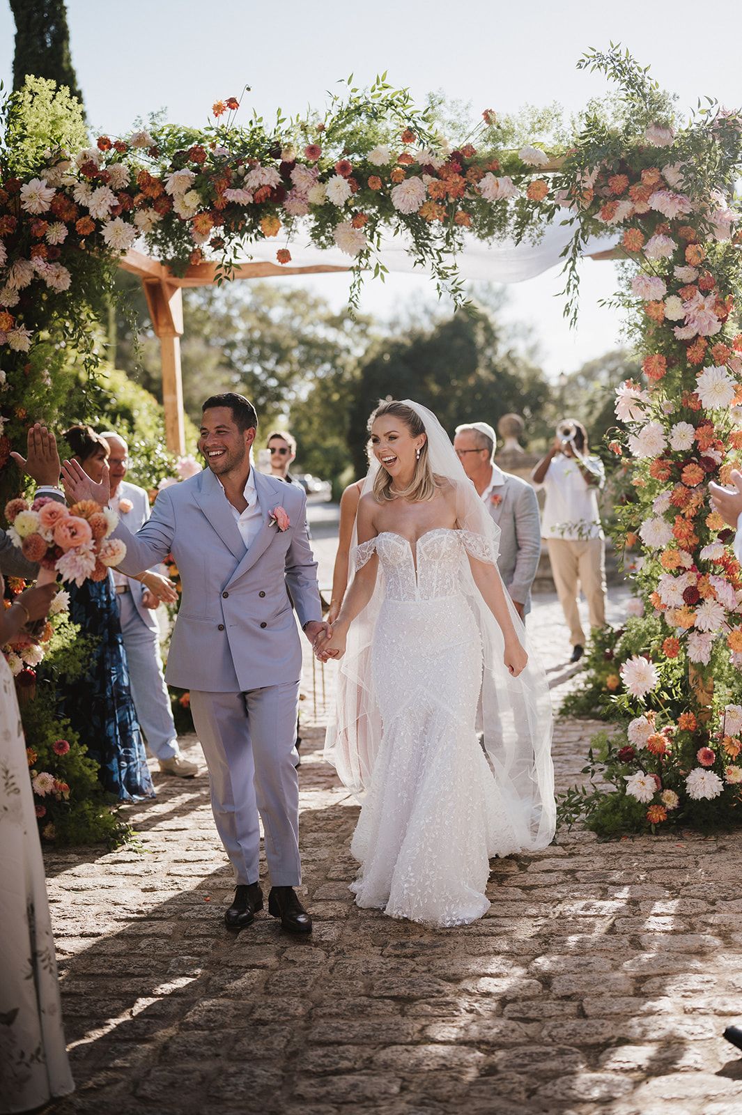 Joyful bride and groom exit under floral pergola arch lined with peach and pink blooms at outdoor wedding.