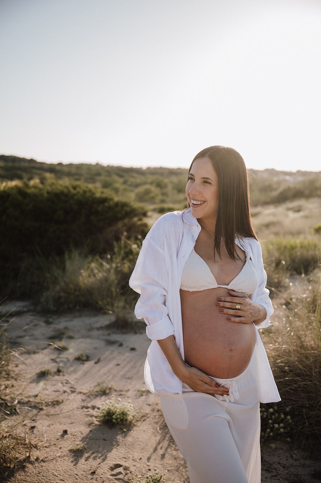 Smiling pregnant woman in white outfit holding baby bump in sunny outdoor field setting.