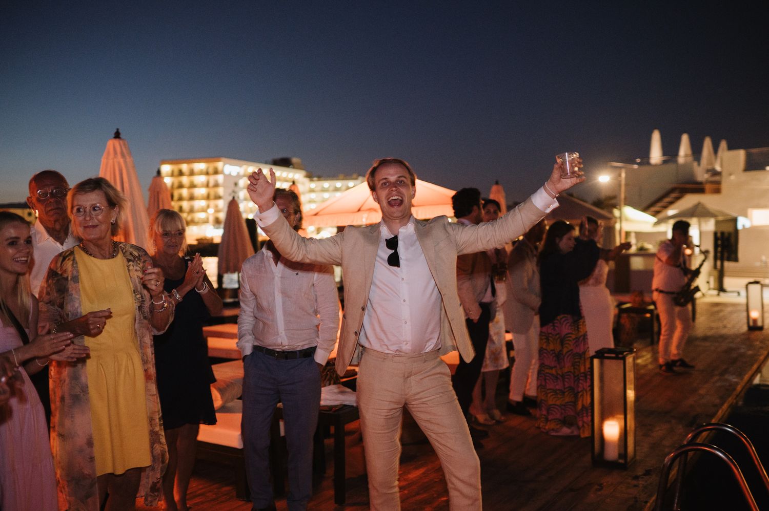 Man in white suit celebrates with arms raised on dance floor at outdoor evening wedding reception.