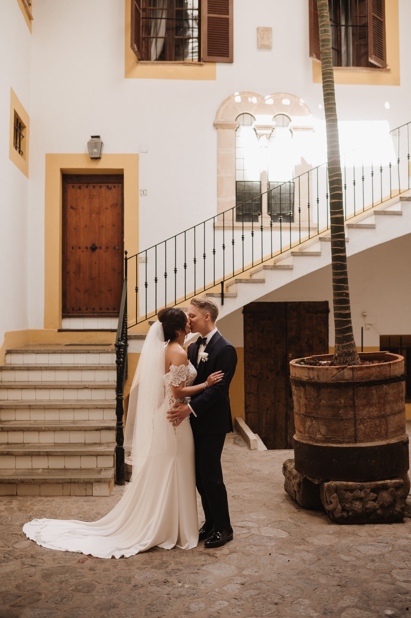 Bride and groom embrace in an elegant courtyard with stone staircase and palm tree.