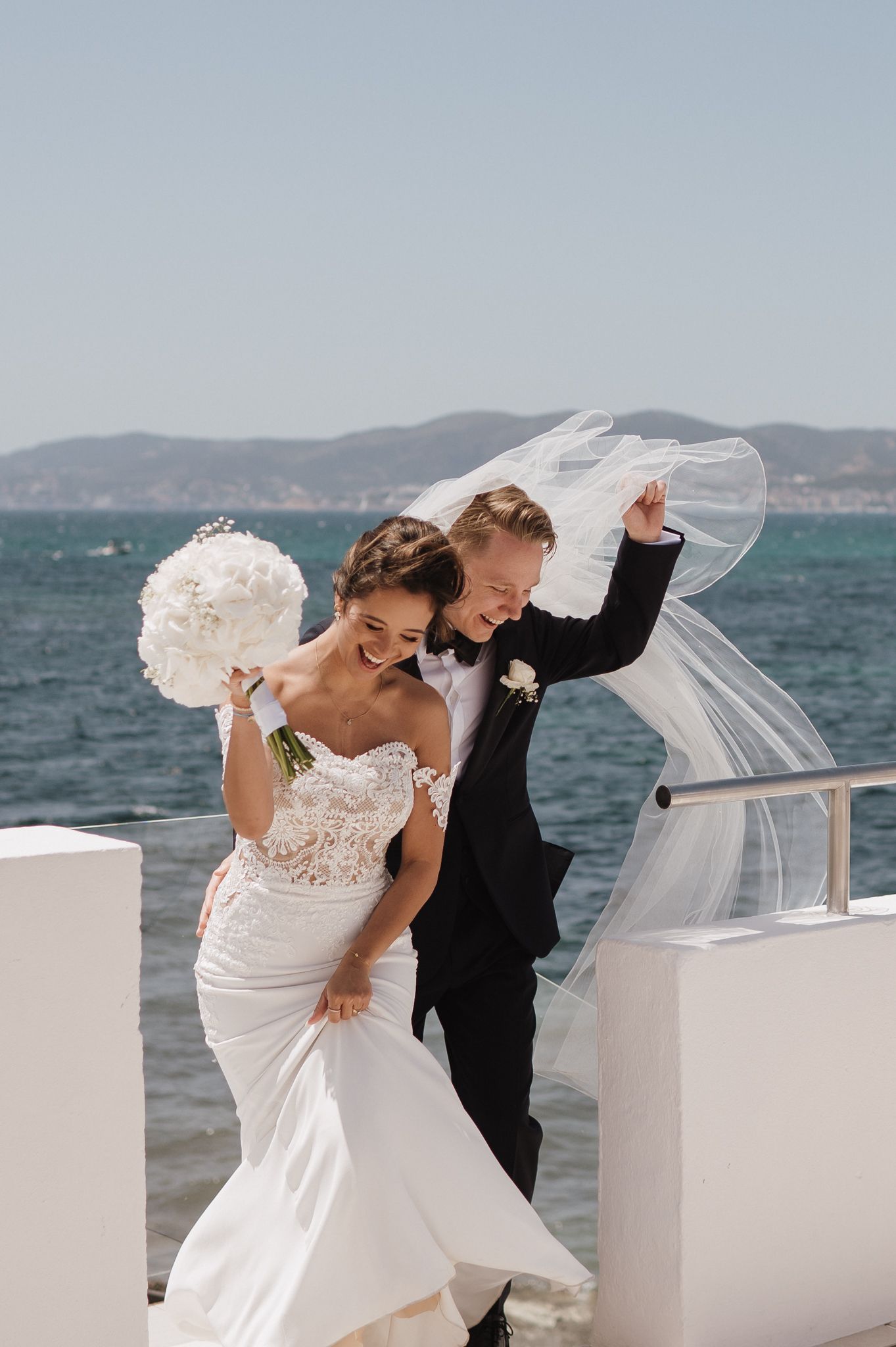 Joyful newlyweds laugh as bride's veil blows in wind by the sea, bride holding white bouquet.