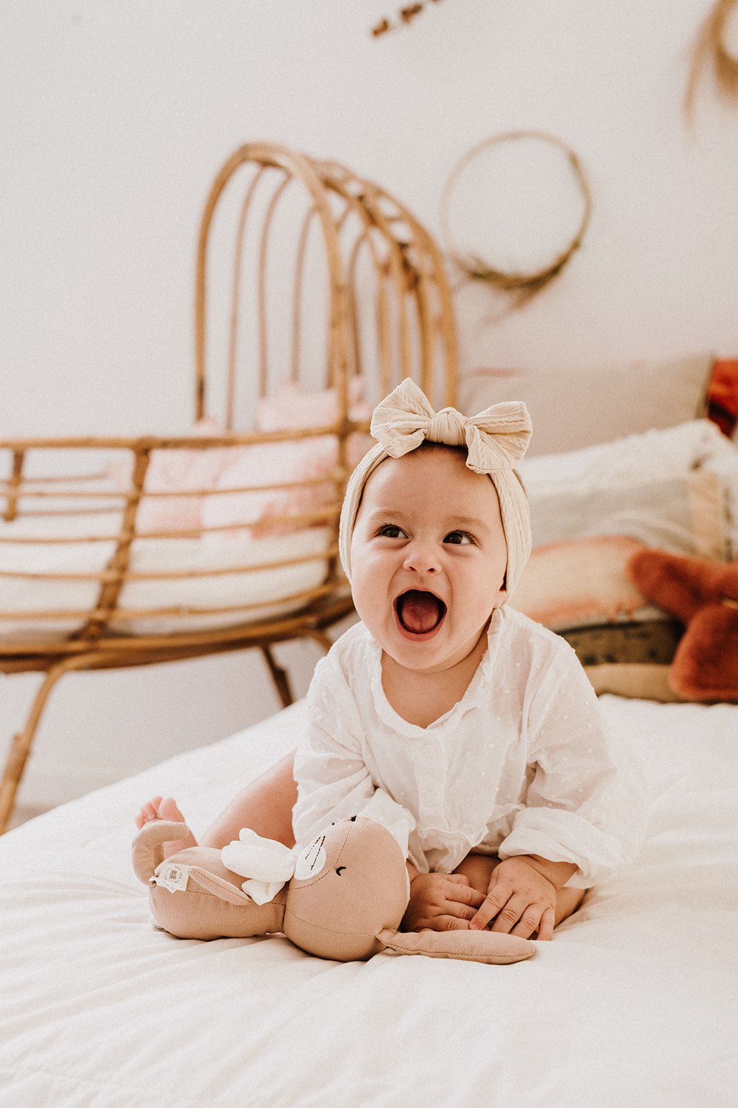Adorable baby girl in white dress and bow headband sitting on bed with stuffed toy, mouth open wide.