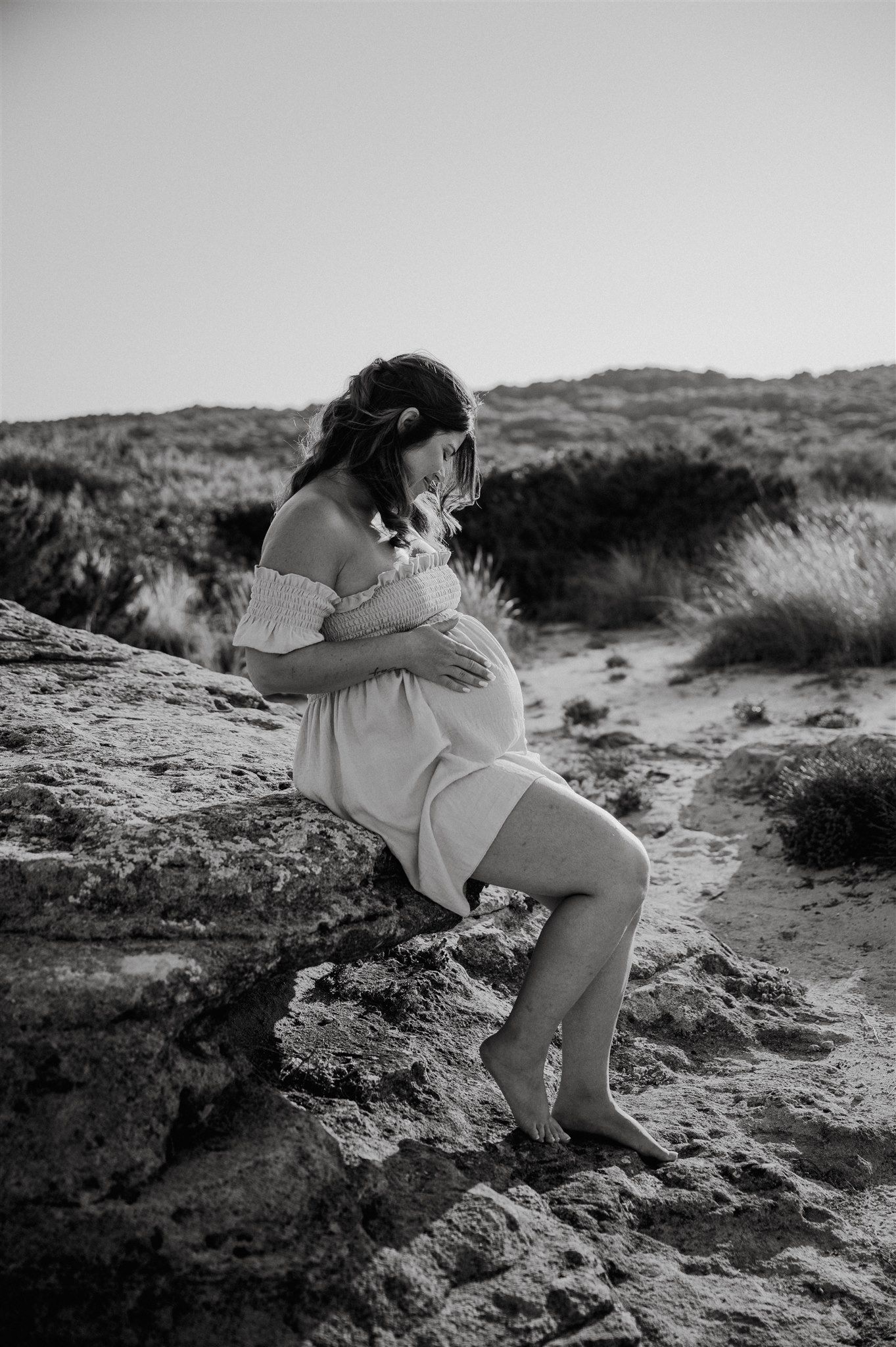Black and white photo of a pregnant woman in a white dress sitting on rocks at a sandy beach, cradling her belly.